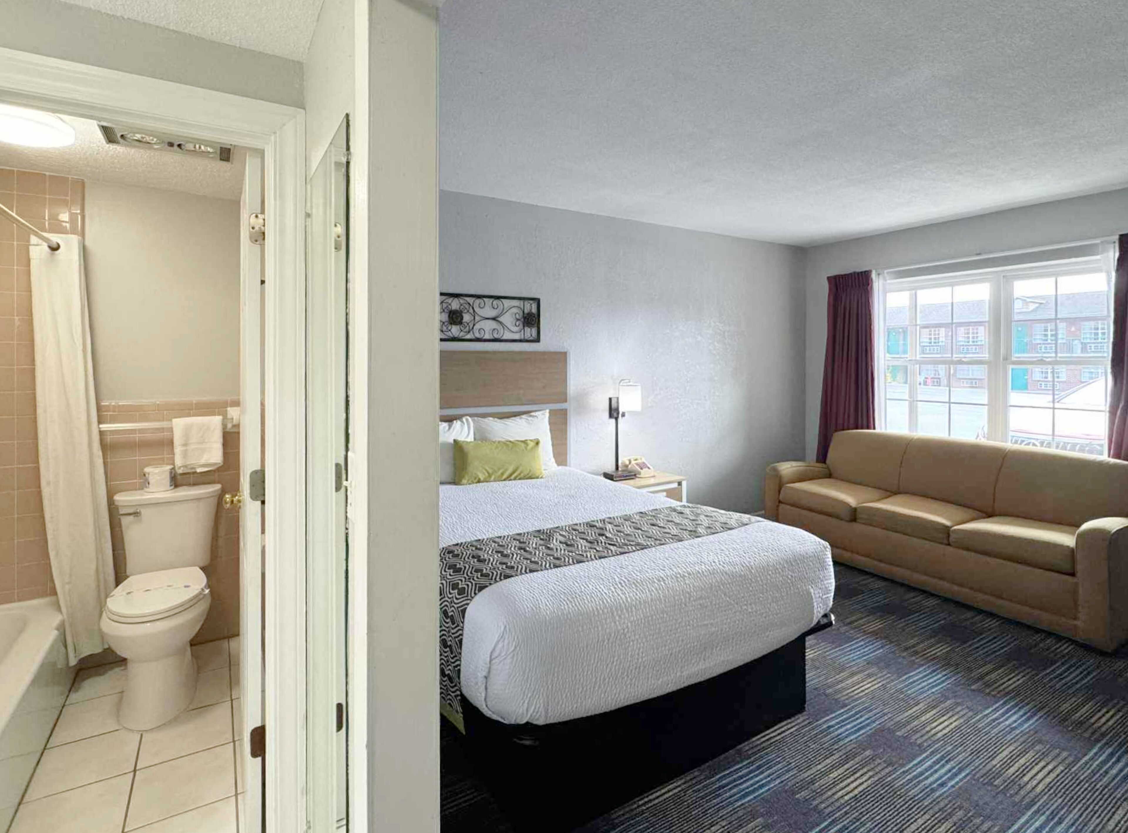 Habitación de hotel con baño, cama con colcha estampada, mesita de noche con lámpara y sofá.