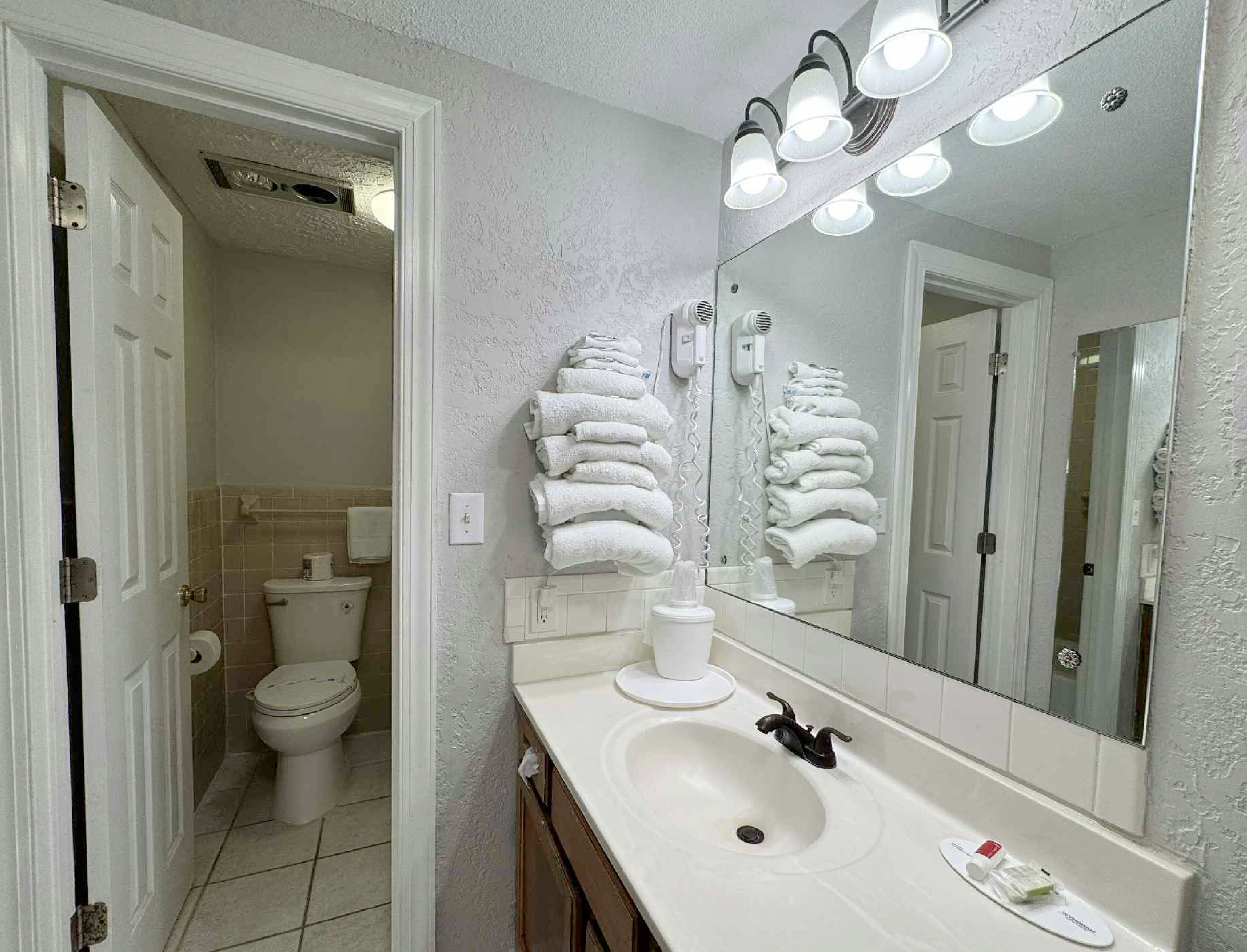 A bathroom with a white sink, a mirror with lights, and a stack of white towels on the wall.