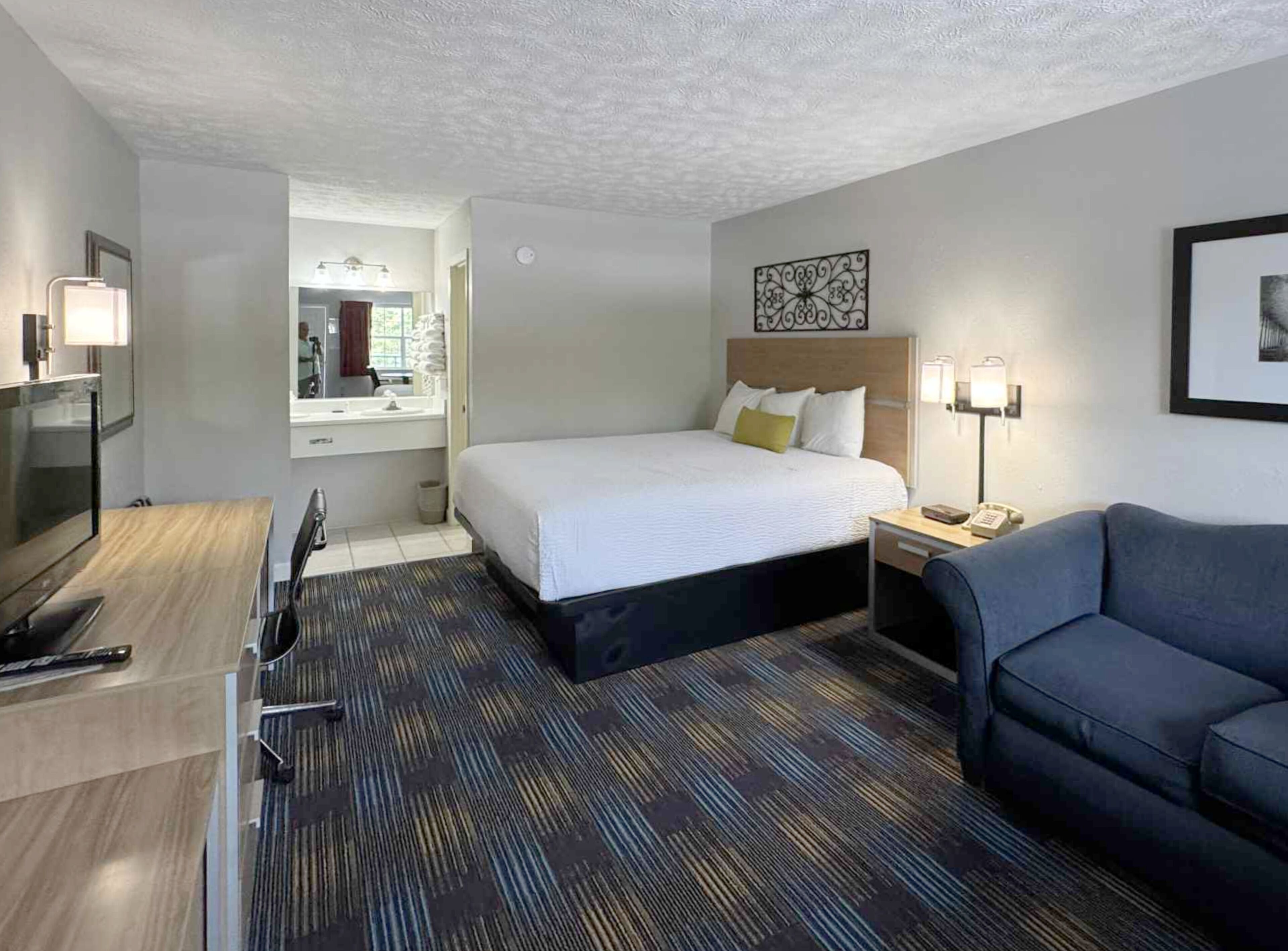 A hotel room with a bed, a desk, a chair, a sofa, and a bathroom with a mirror and sink.