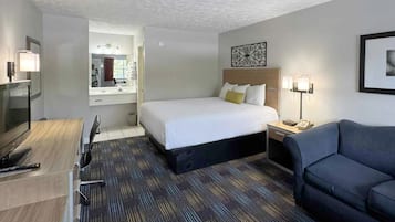 A hotel room with a bed, a desk, a chair, a sofa, and a bathroom with a mirror and sink.