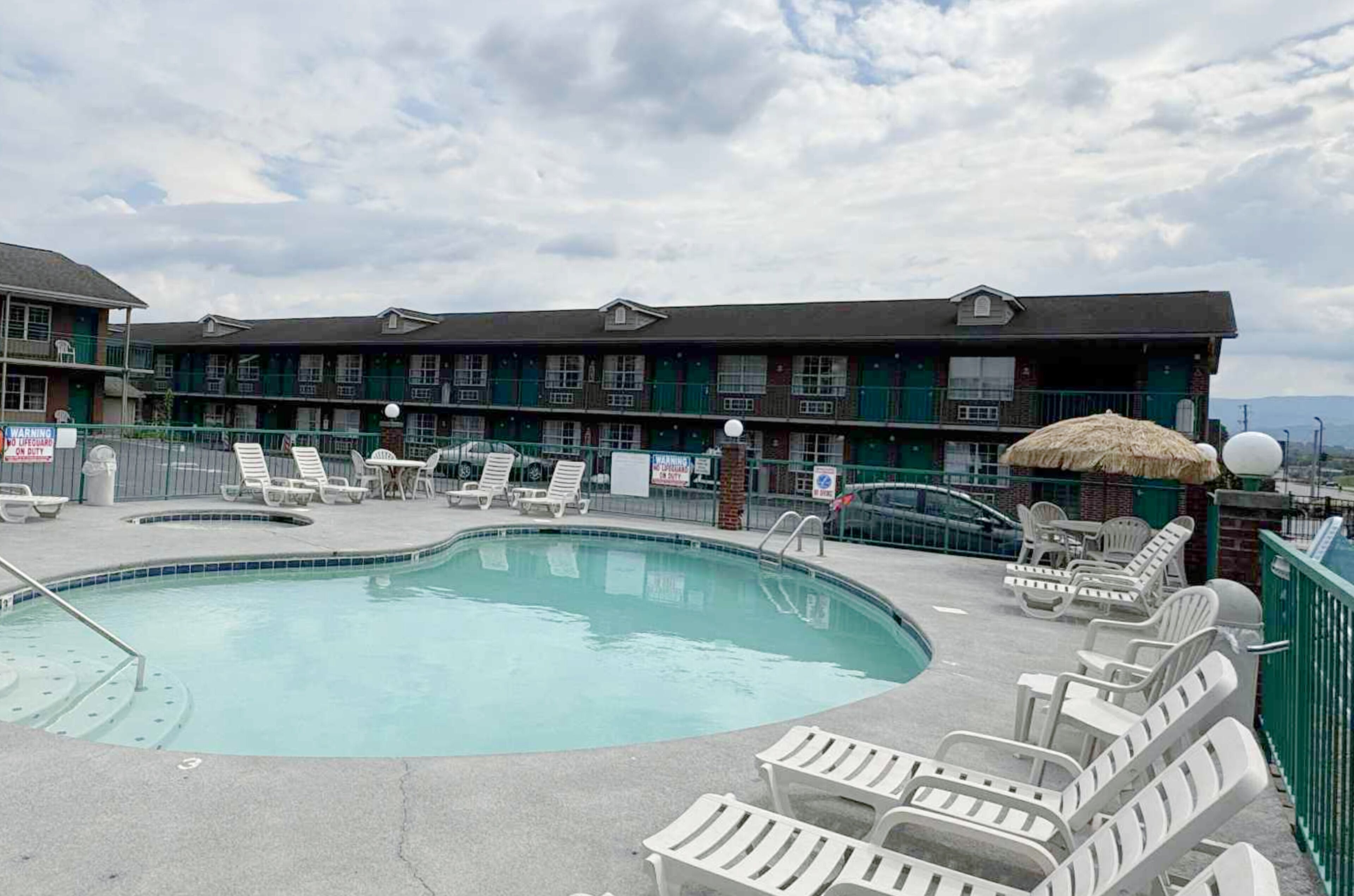A motel with a swimming pool, lounge chairs, and a thatched umbrella.