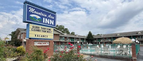 A Mountain Aire Inn sign with a clear view of the motel's exterior, including a brick building, a covered parking area, and a garden with red flowers.