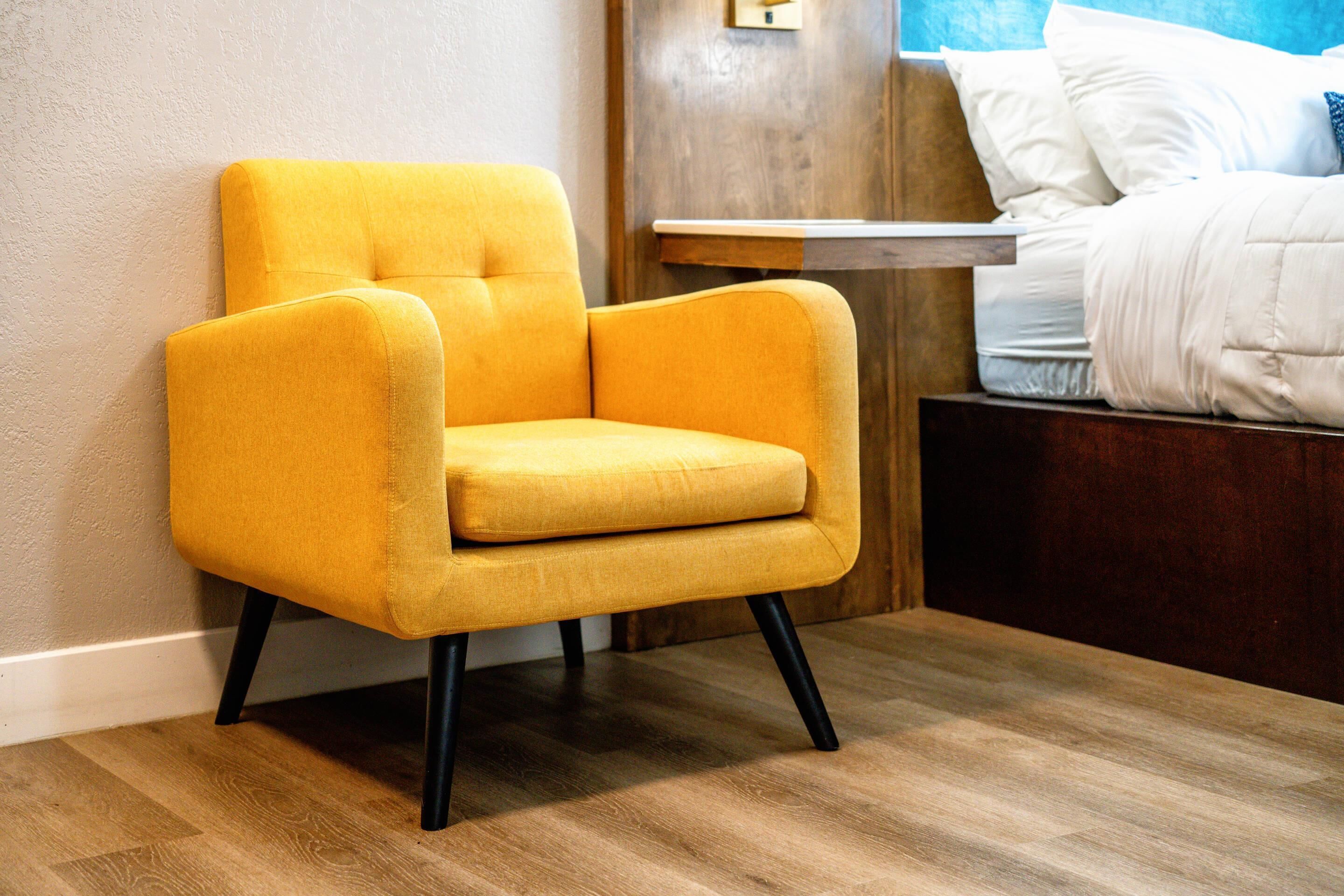 A yellow armchair with black legs, a wooden floor, a bedside table, and a bed with white bedding.