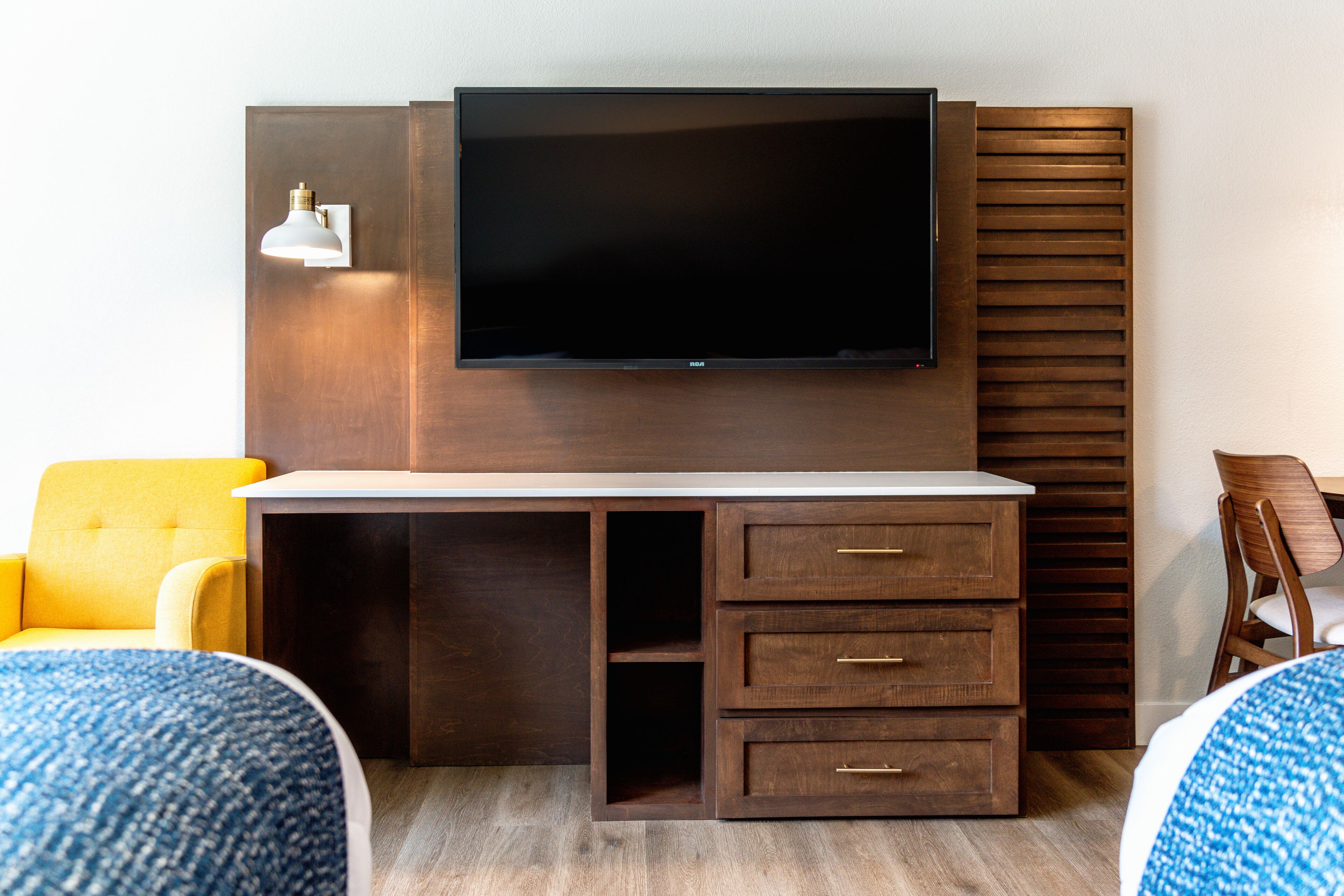 A hotel room with a flat-screen TV mounted on a wooden wall, a small desk with drawers, a yellow armchair, and a blue patterned ottoman.