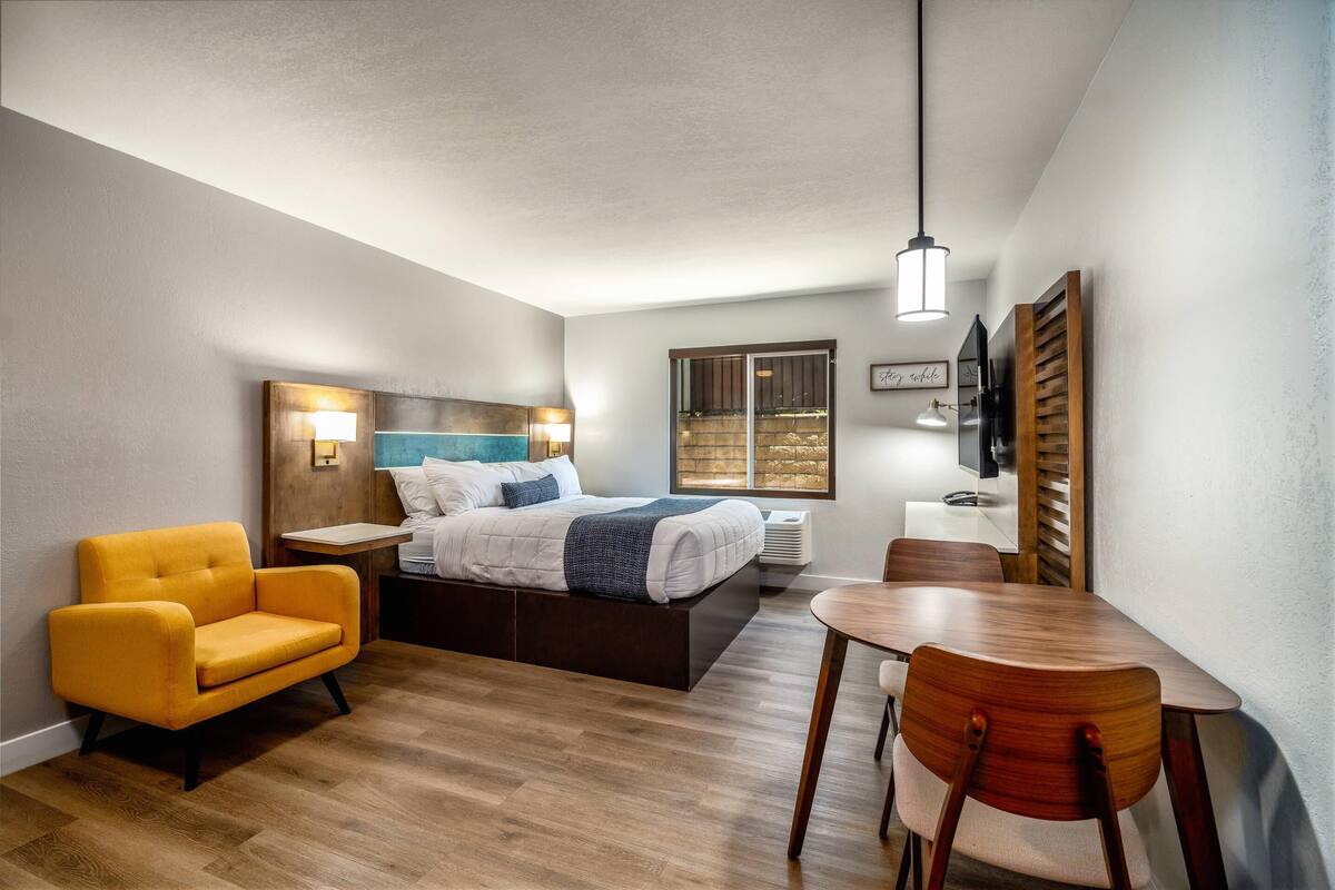 A modern hotel room with a bed, a yellow armchair, a wooden dining table, and a small side table.