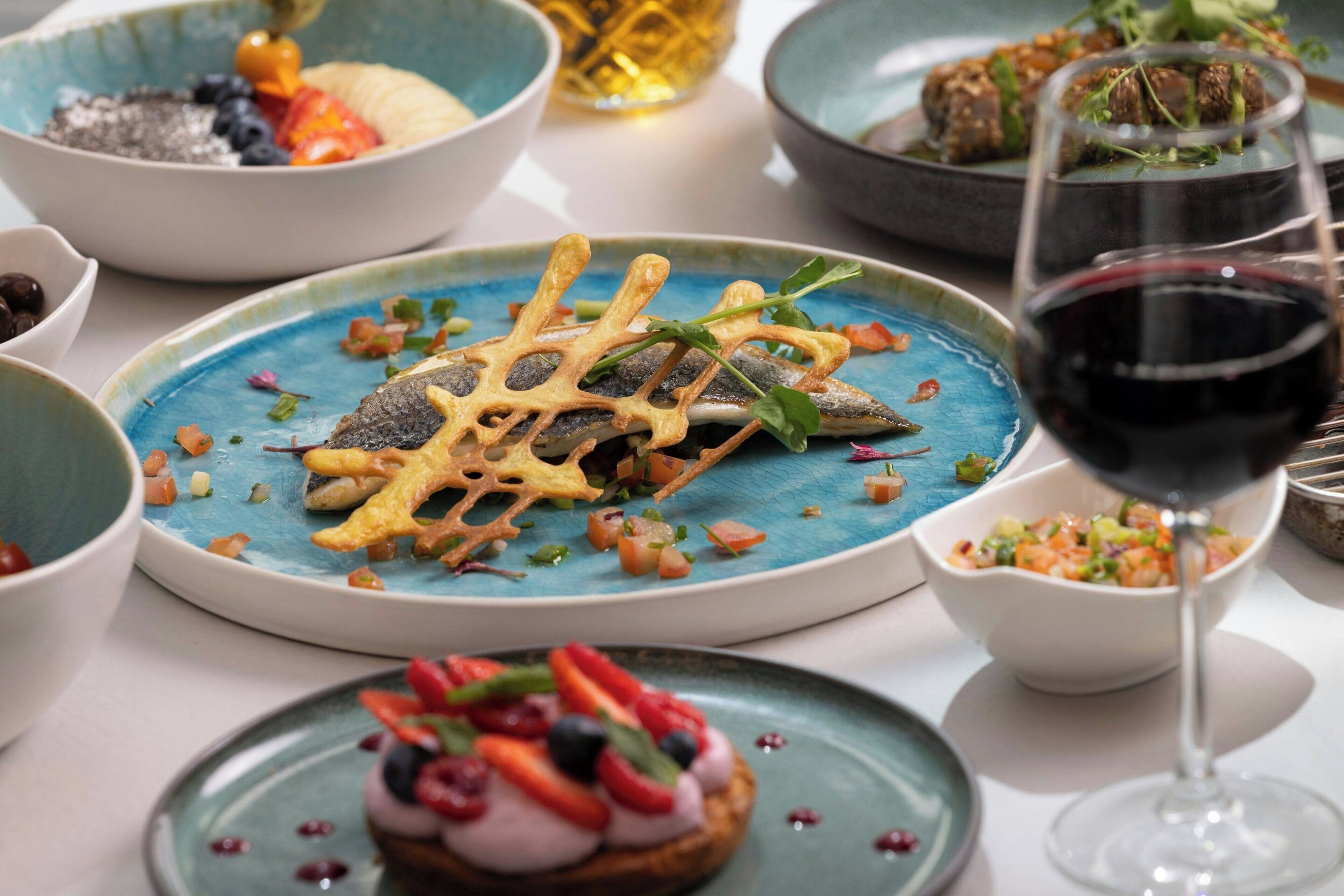 A variety of dishes on a table, including a fish with a decorative lattice on top, a glass of red wine, and several bowls with different food items.