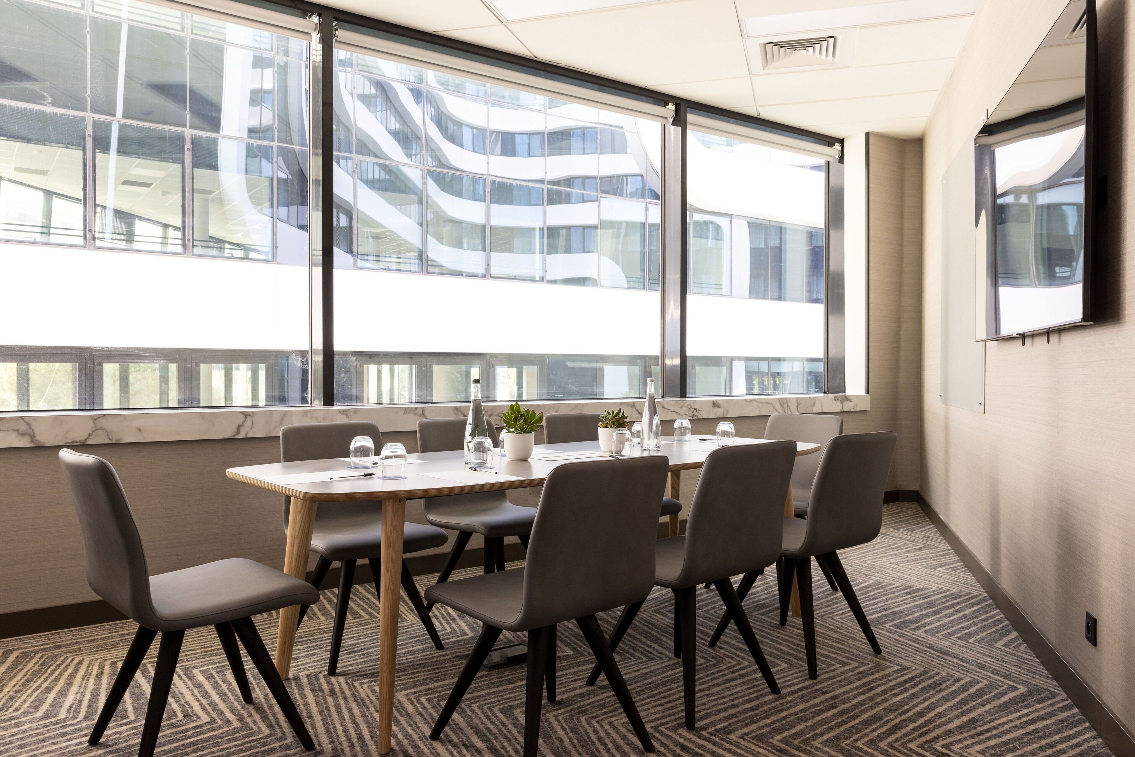Uma sala de conferências com uma mesa grande, seis cadeiras e vista para um edifício moderno com janelas de vidro.