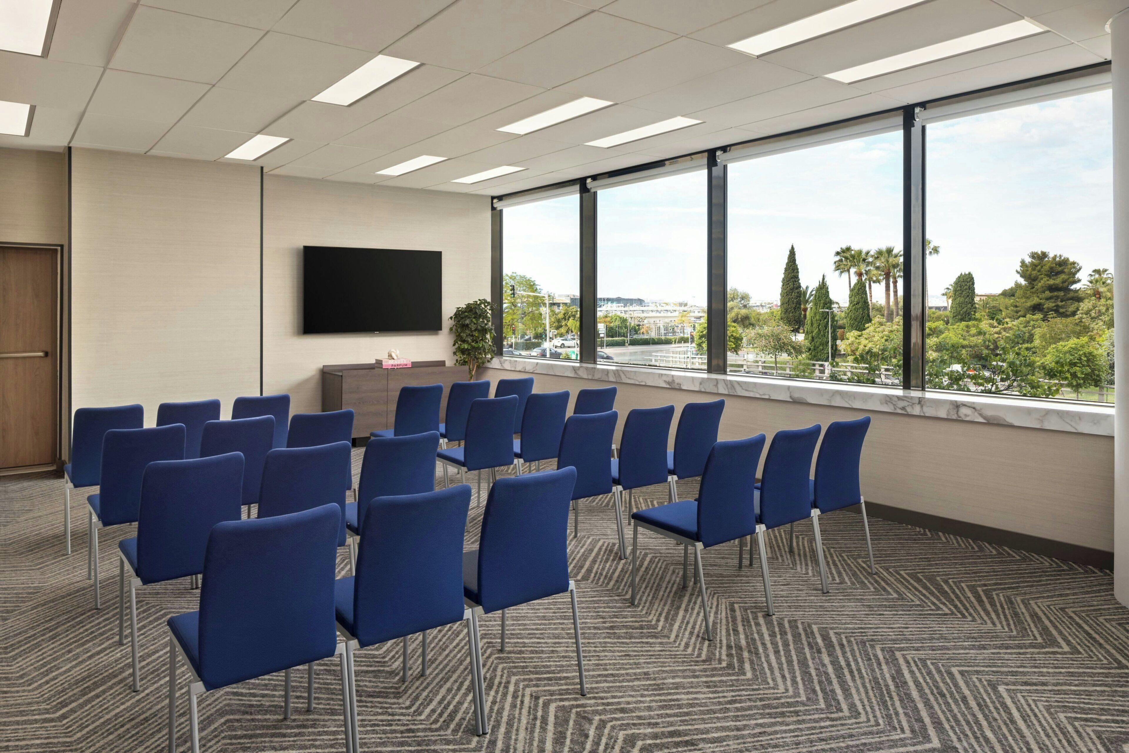 A conference room with rows of chairs, a flat-screen TV on the wall, and a large window with a view of trees and buildings.
