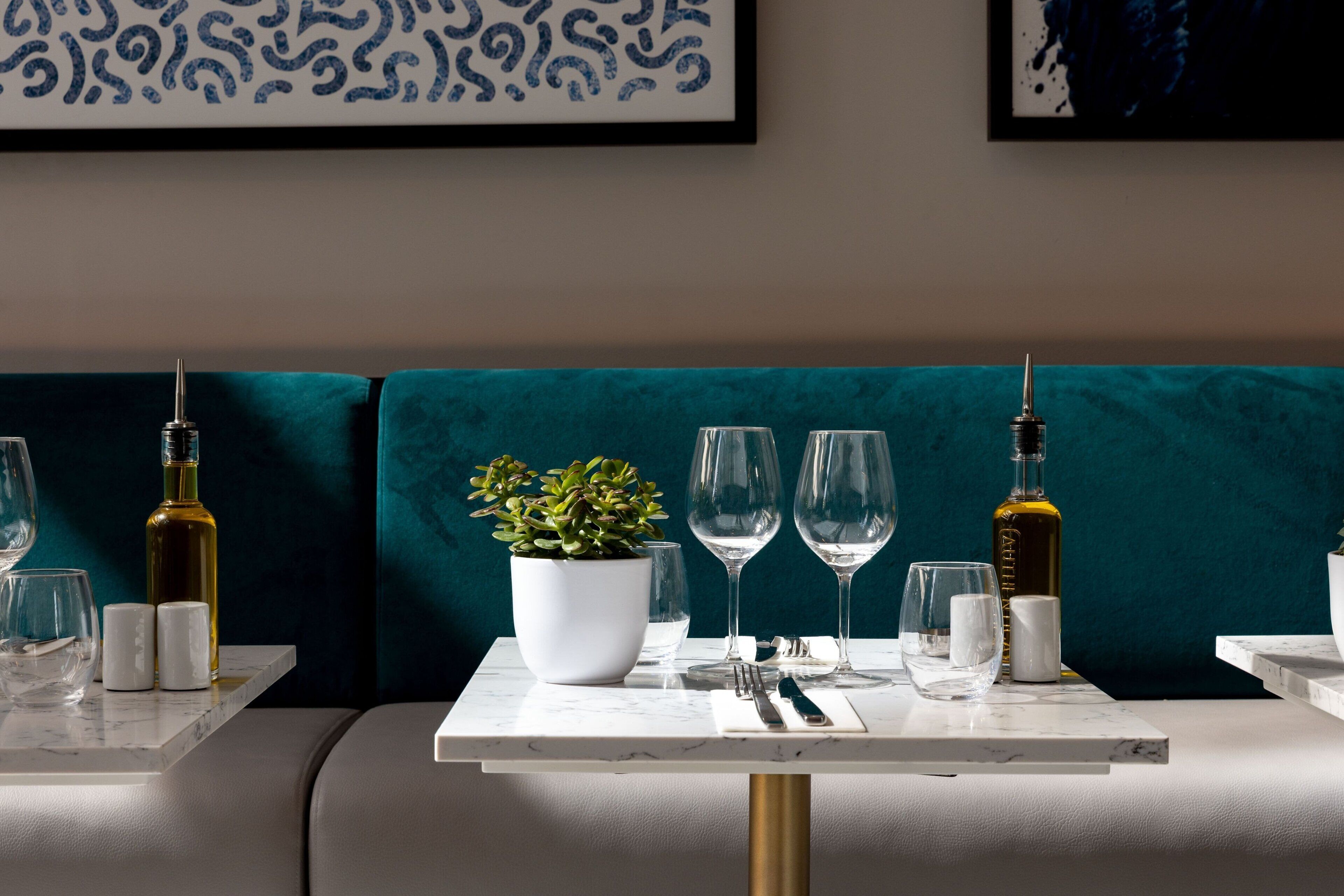Un área de comedor con un banco tapizado en color turquesa, una mesa blanca con cubierta de mármol, una planta en maceta, copas de vino y botellas.