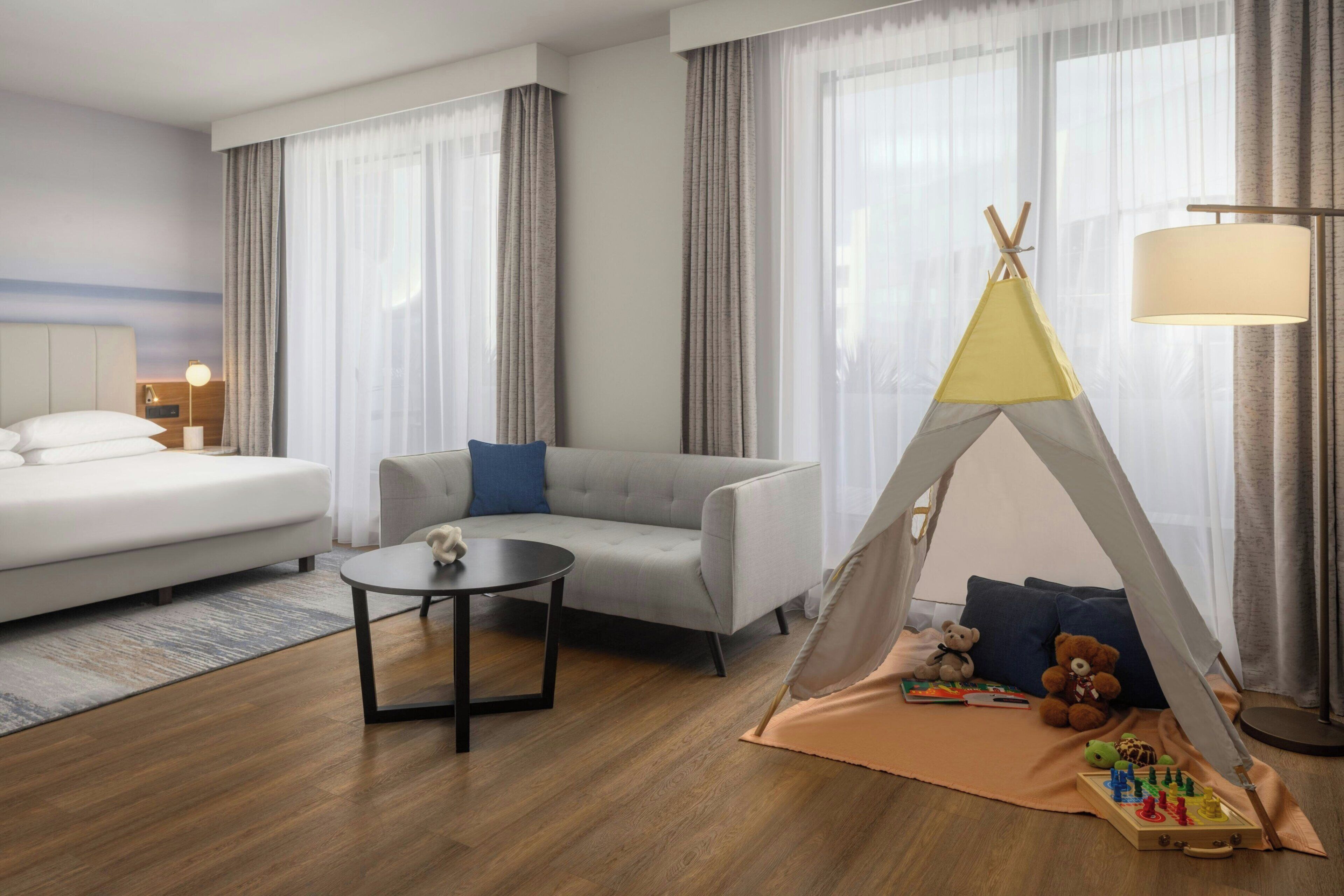 Une chambre d’hôtel avec un lit, un canapé, une petite table, une lampe, une tente et des jouets pour enfants.