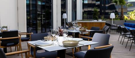 An outdoor dining area with tables and chairs, a counter, and a view of a modern building with large windows.