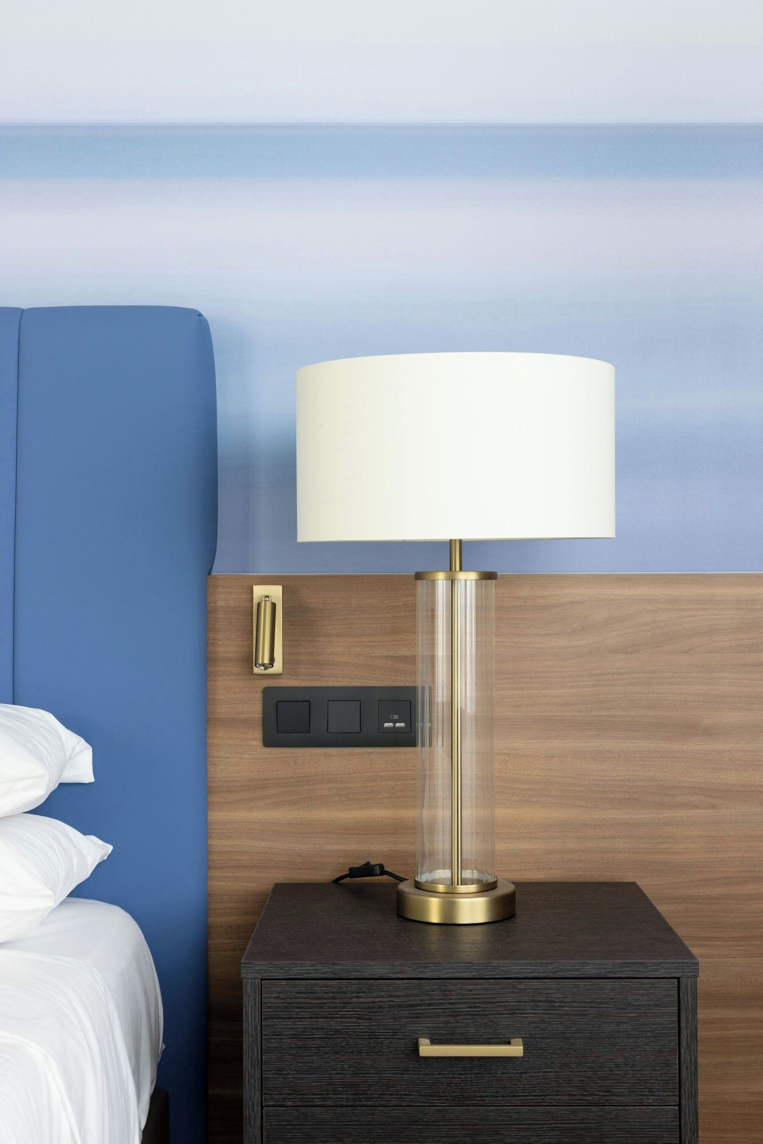 A hotel room with a bed, a nightstand with a lamp, a control panel, and a view of the ocean.