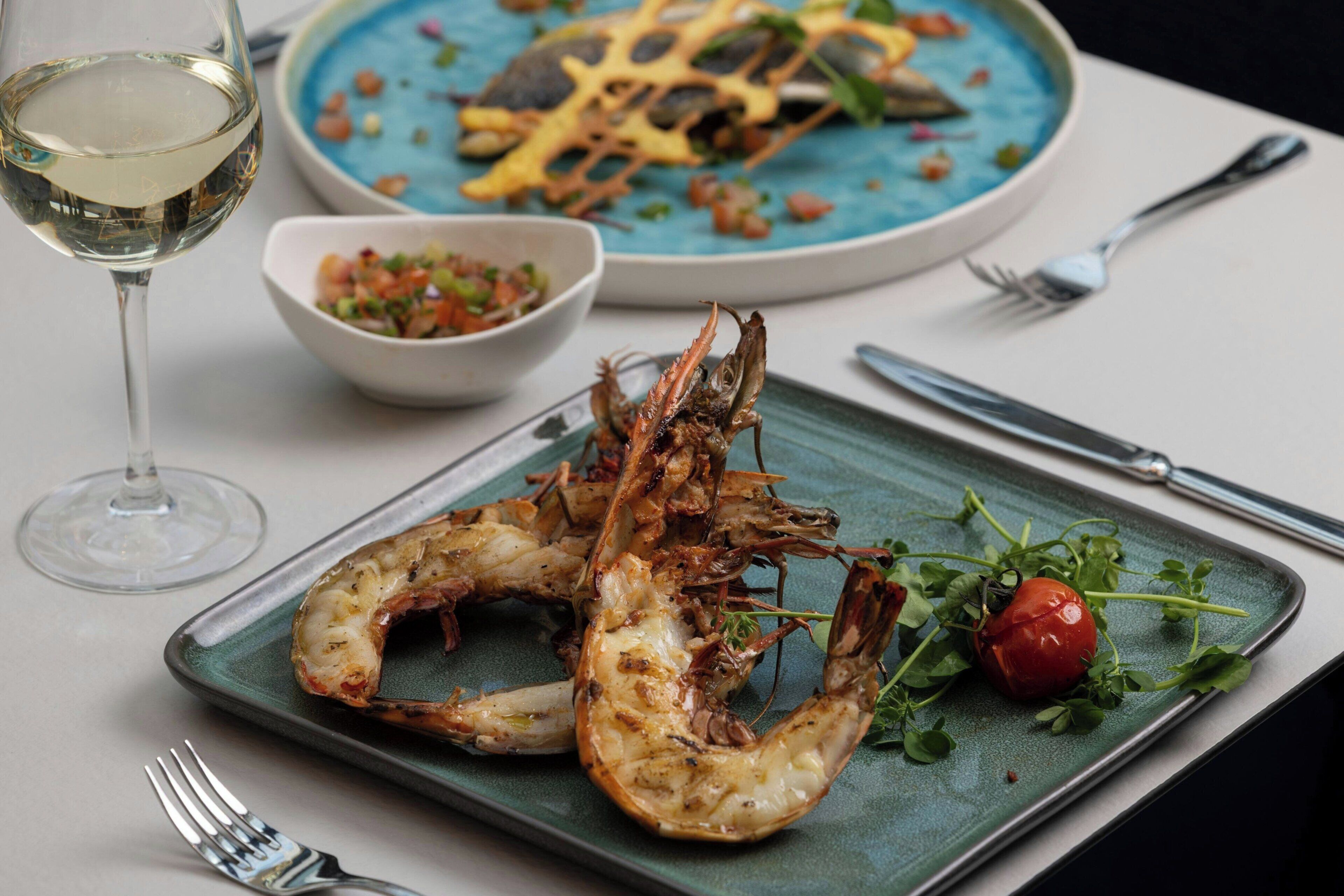 A plate of grilled shrimp with a garnish of greens and a cherry tomato, a glass of white wine, and a bowl of salad on a table.