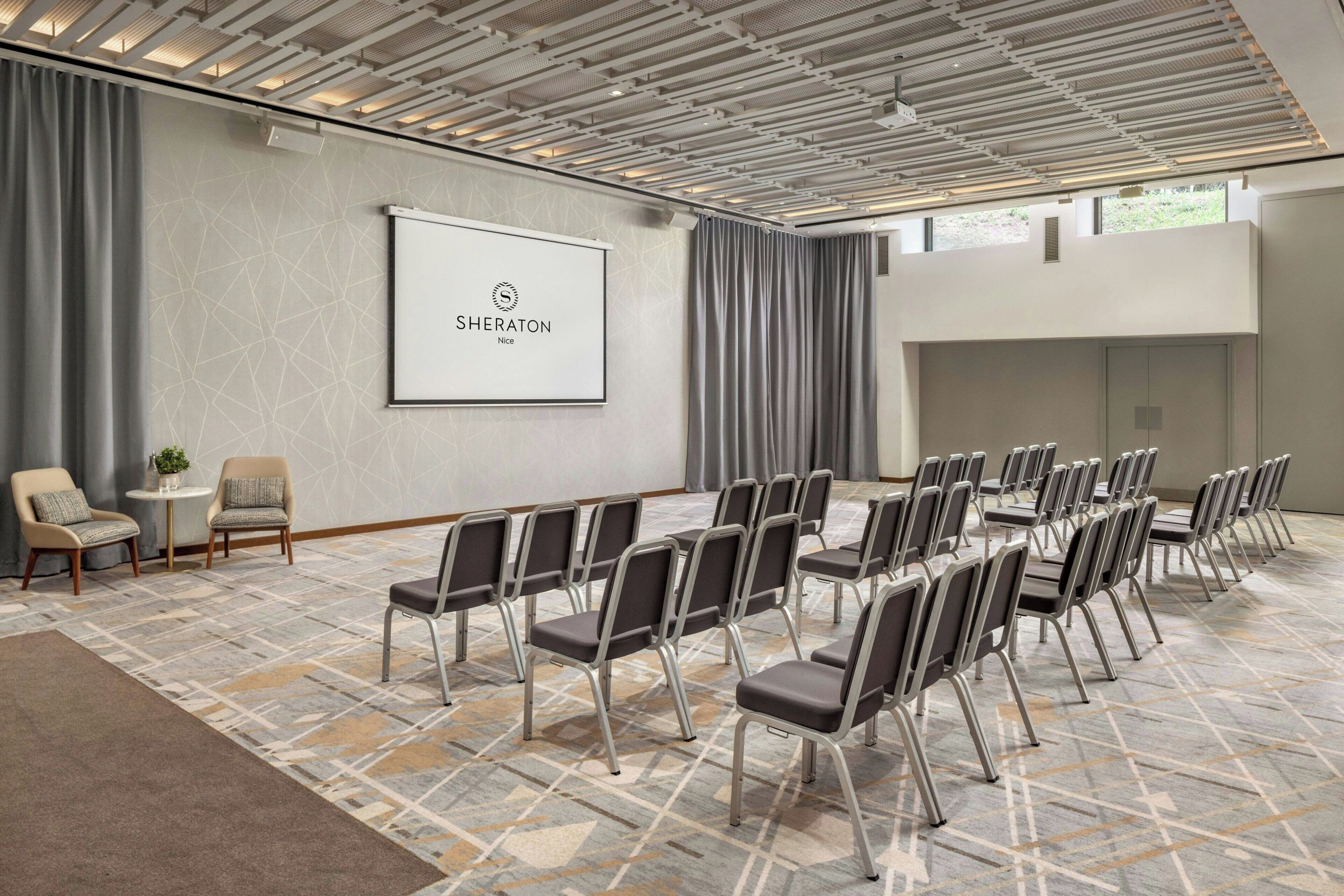 Une salle de conférence équipée d’un écran de projection affichant le logo « Sheraton Nice », avec des sièges disposés de manière ordonnée et une moquette à motifs.