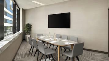 Un salón de conferencias con una larga mesa, seis sillas, una planta en maceta y una gran pantalla negra en la pared.