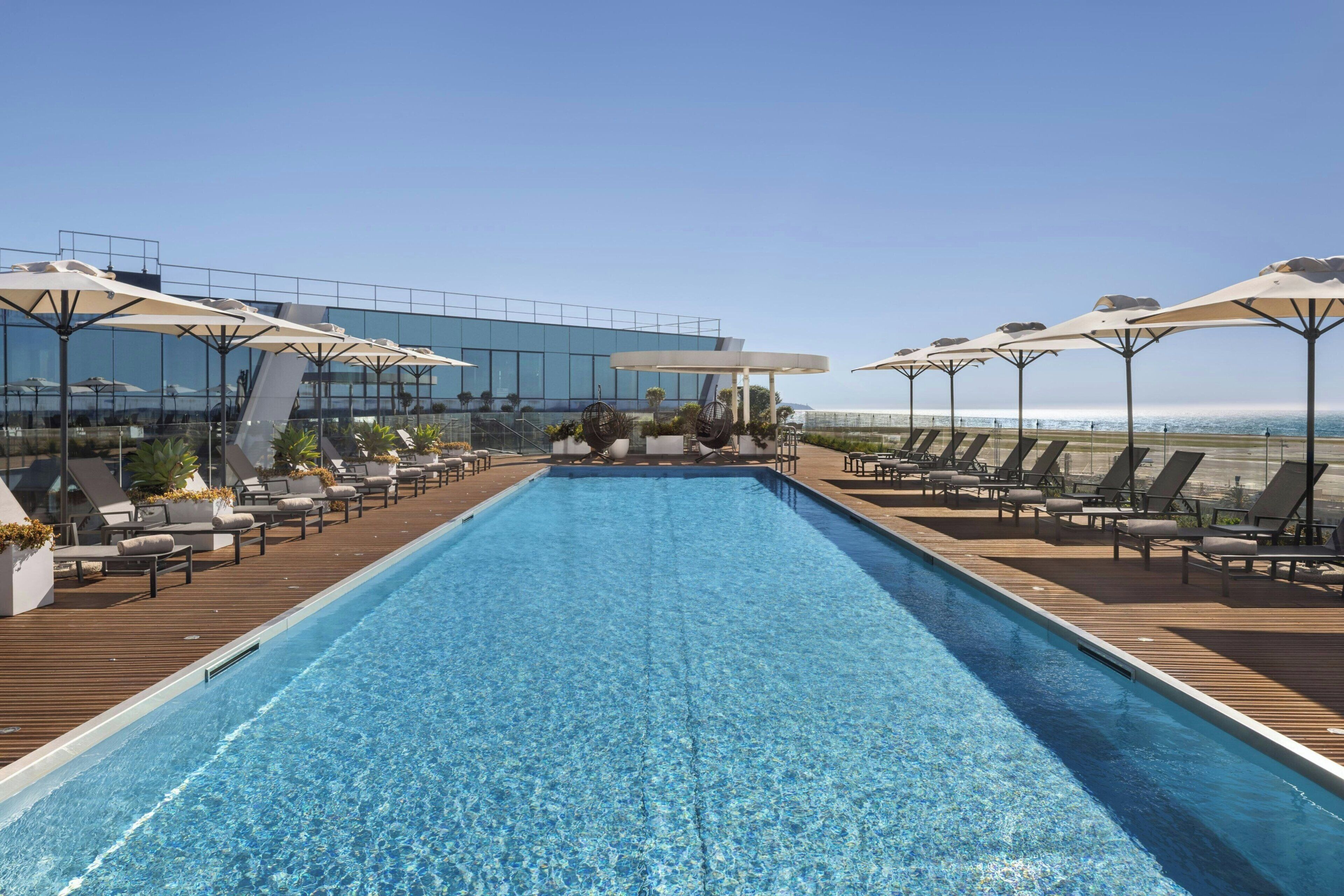 Piscina infinita en la azotea con sillones de descanso y sombrillas, con vistas a un paisaje urbano.