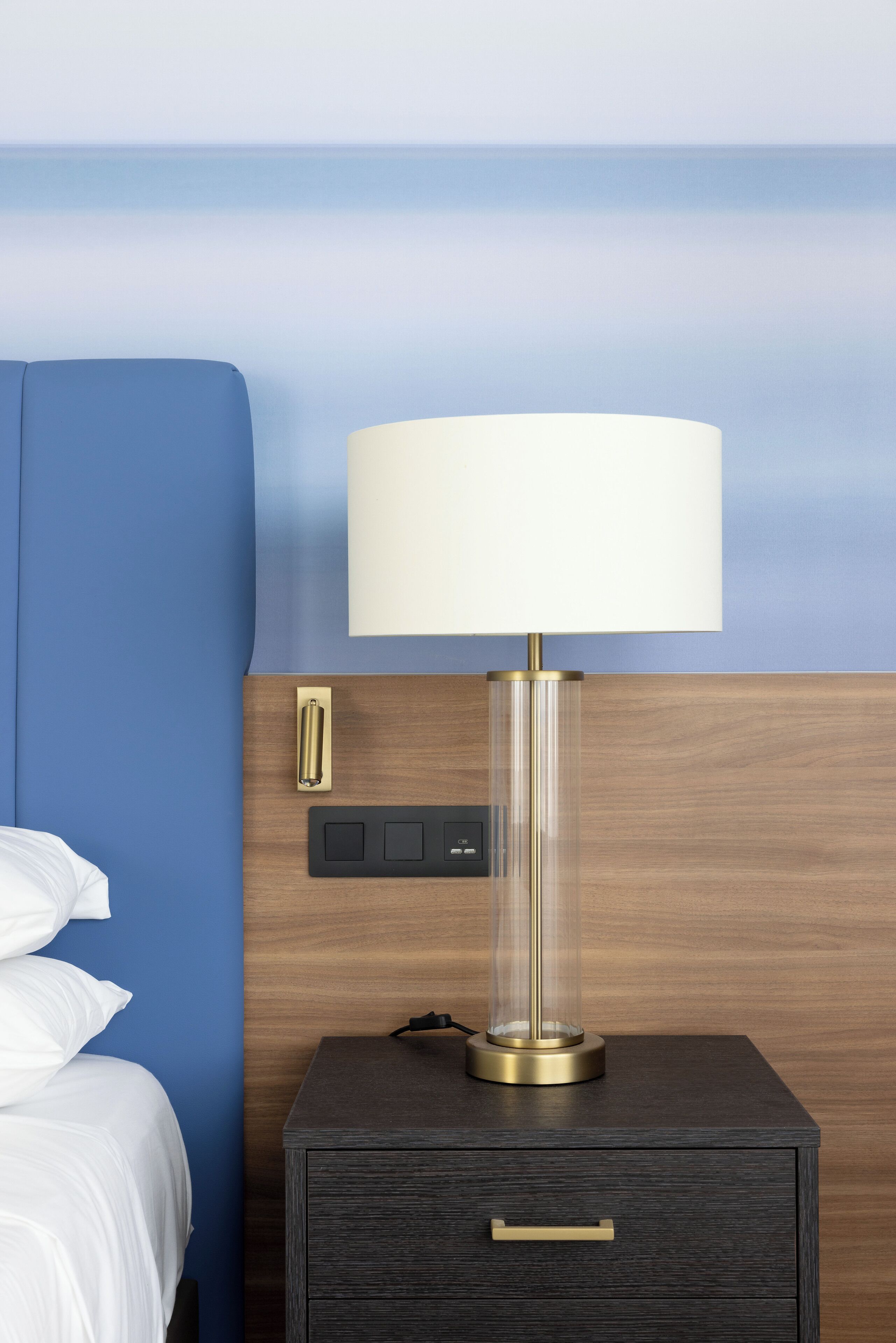 Une chambre d’hôtel comprenant un lit, une table de chevet avec une lampe, une prise de courant et un panneau de commande.