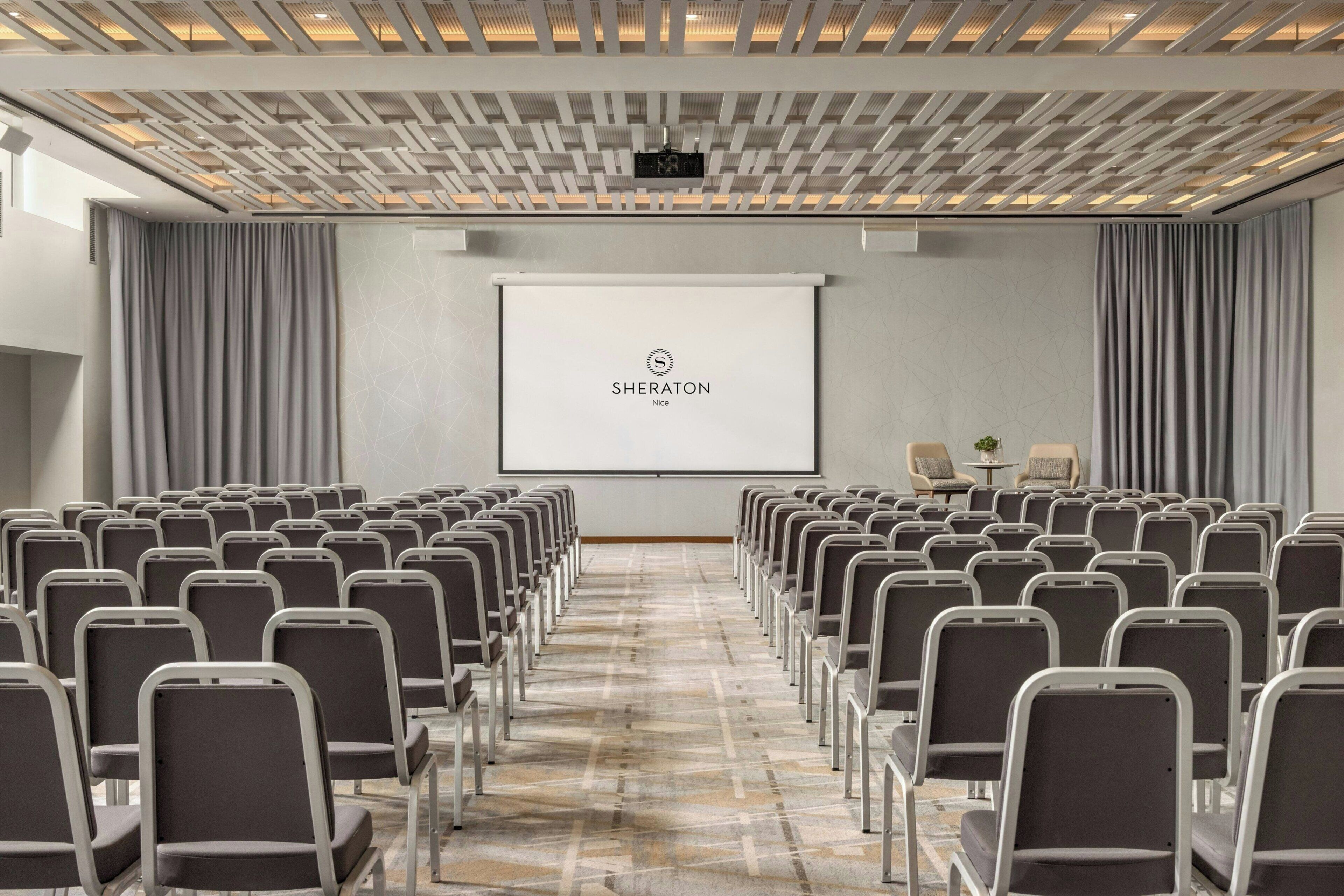 Una sala de conferències amb files de cadires, una pantalla de projector i un logotip a la pantalla.