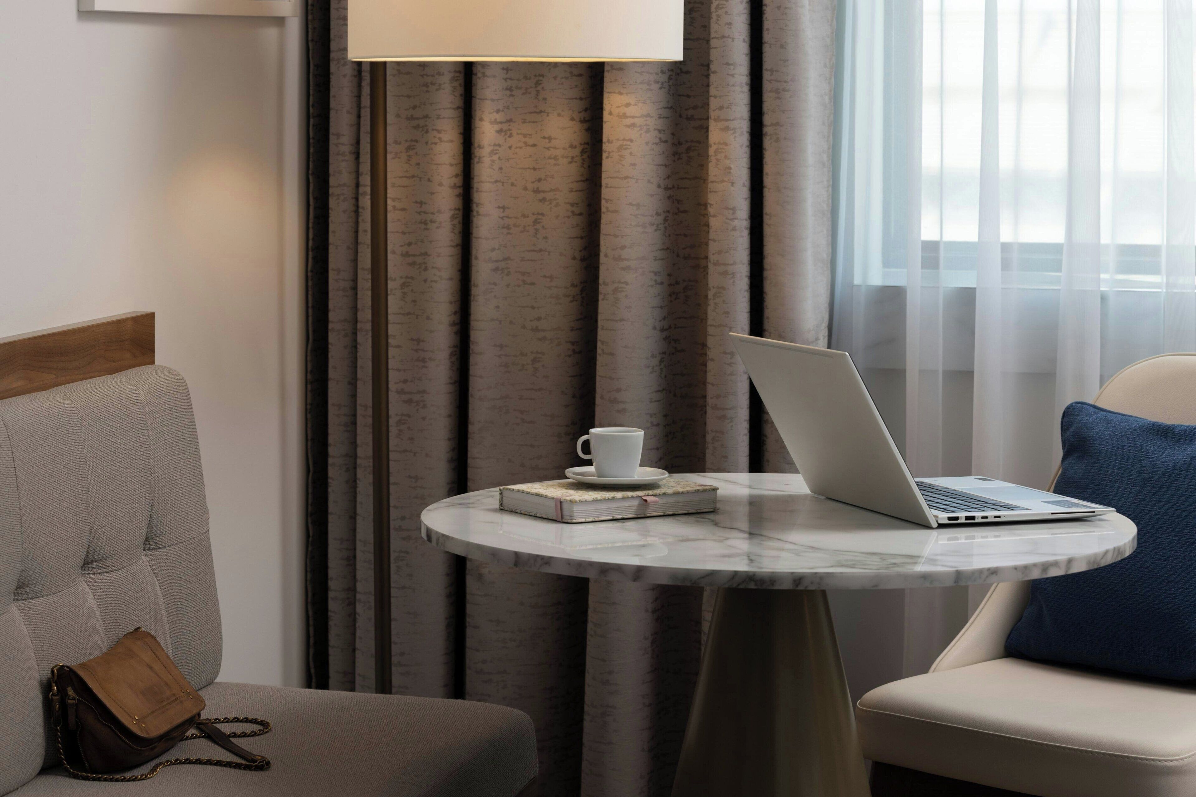 Et hotelværelse med et rundt marmorbord, en bærbar computer, en kop og en bog.