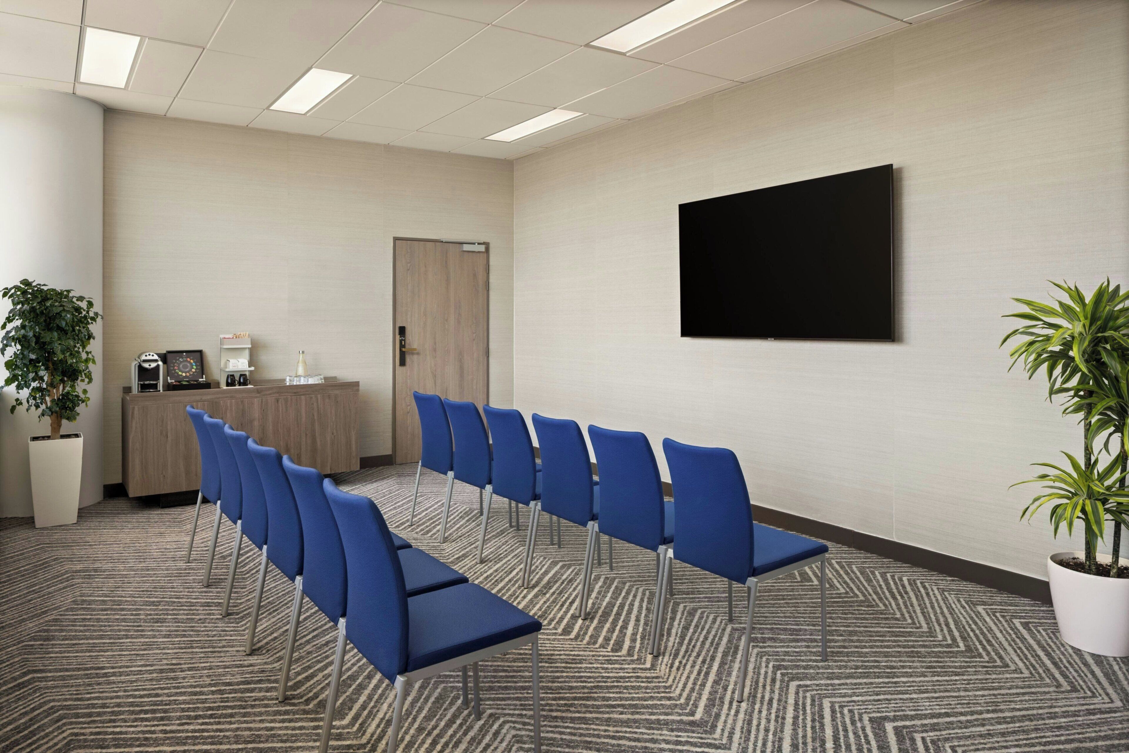 A conference room with a long table, blue chairs, a reception desk, a door, a flat-screen TV, and potted plants.