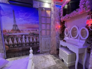 Quarto com um grande mural da Torre Eiffel, uma porta de madeira, uma cômoda com espelhos e um arranjo floral.