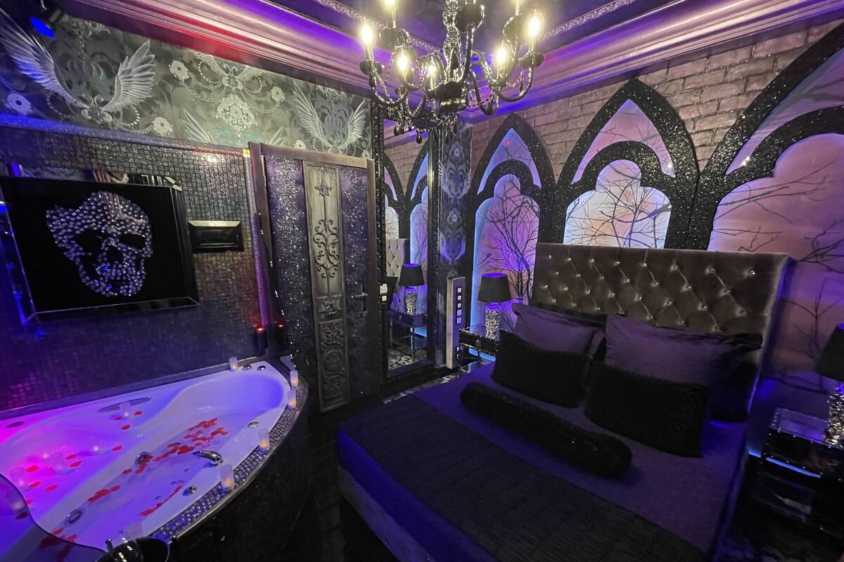 A room with a large bathtub, a bed with a tufted headboard, a chandelier, and gothic-style wall decorations.