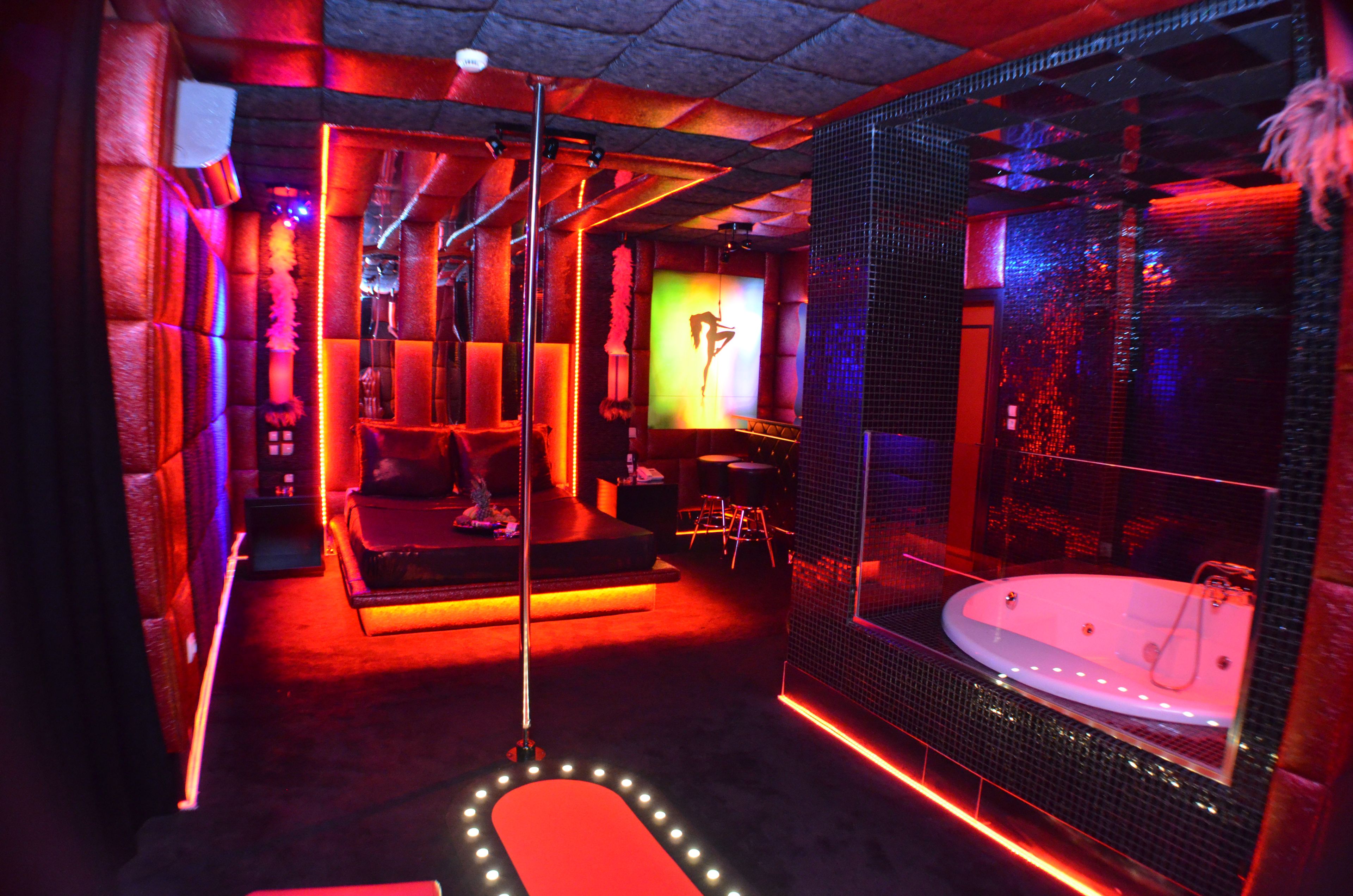 A room with a red-lit jacuzzi, a hanging chair, and a mirror reflecting the room's interior.