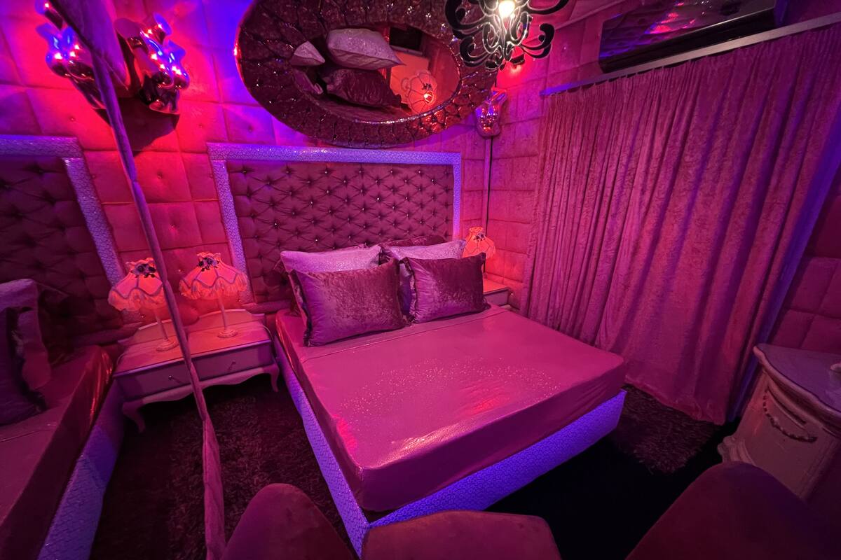 A bedroom with a tufted headboard, a bed with a pink bedspread, a nightstand with a lamp, and a mirror on the wall.