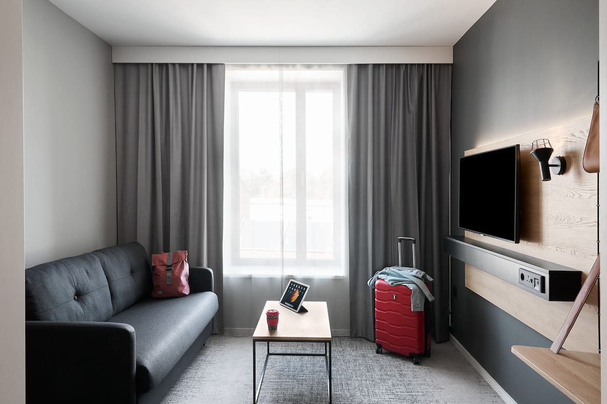 A modern hotel room with a grey sofa, a small table with a tablet and a mug, a red suitcase, and a TV mounted on the wall.