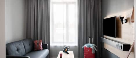 Una habitación de hotel moderna con un sofá gris, una pequeña mesa con una tableta y una taza, una maleta roja y un televisor montado en la pared.