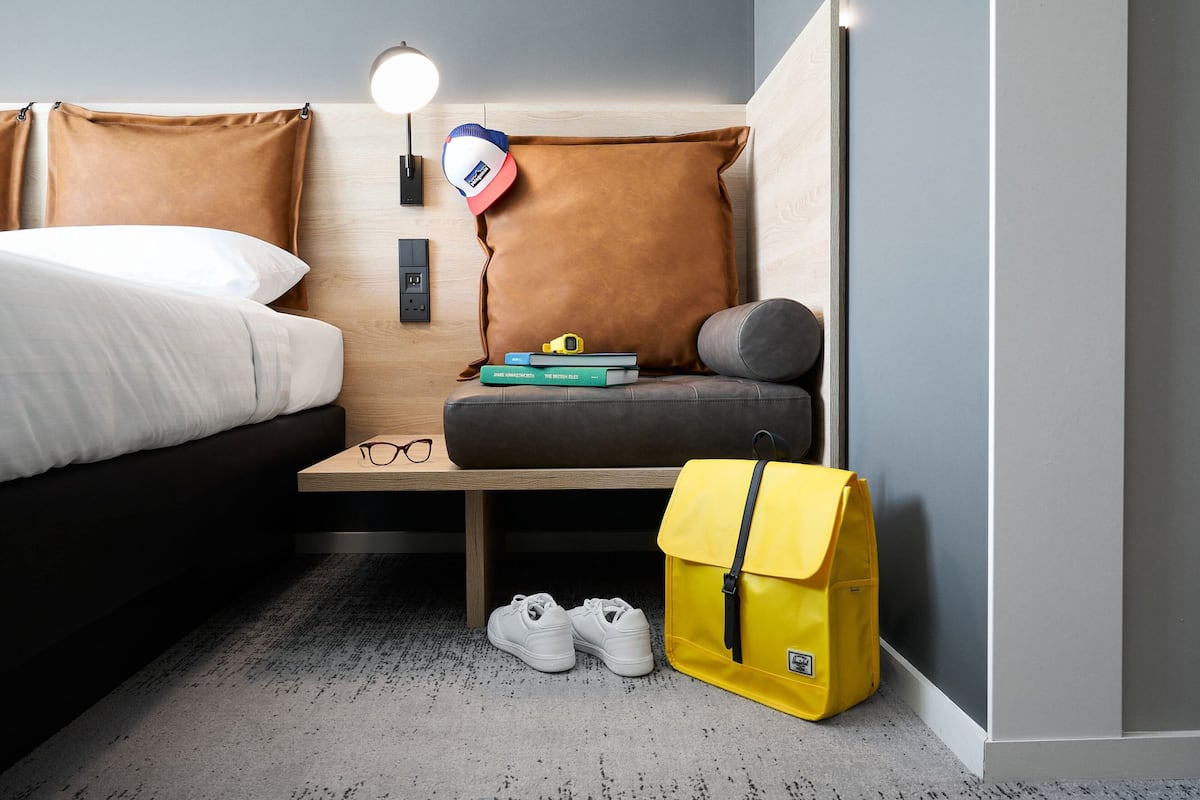 A compact hotel room with a bed, a small desk, a lamp, and a yellow bag on the floor.