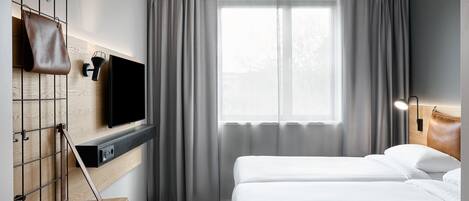 A hotel room with a bed, a TV mounted on a wooden panel, a small table, and a window with curtains.