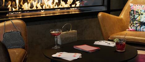 A cozy seating area with a leather couch, a small round table with playing cards and a drink, and a fireplace in the background.