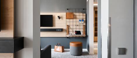 Un interior moderno con alfombra gris, un otoman redond, una silla negra, un estante de madera con un televisor y un aplique de pared.
