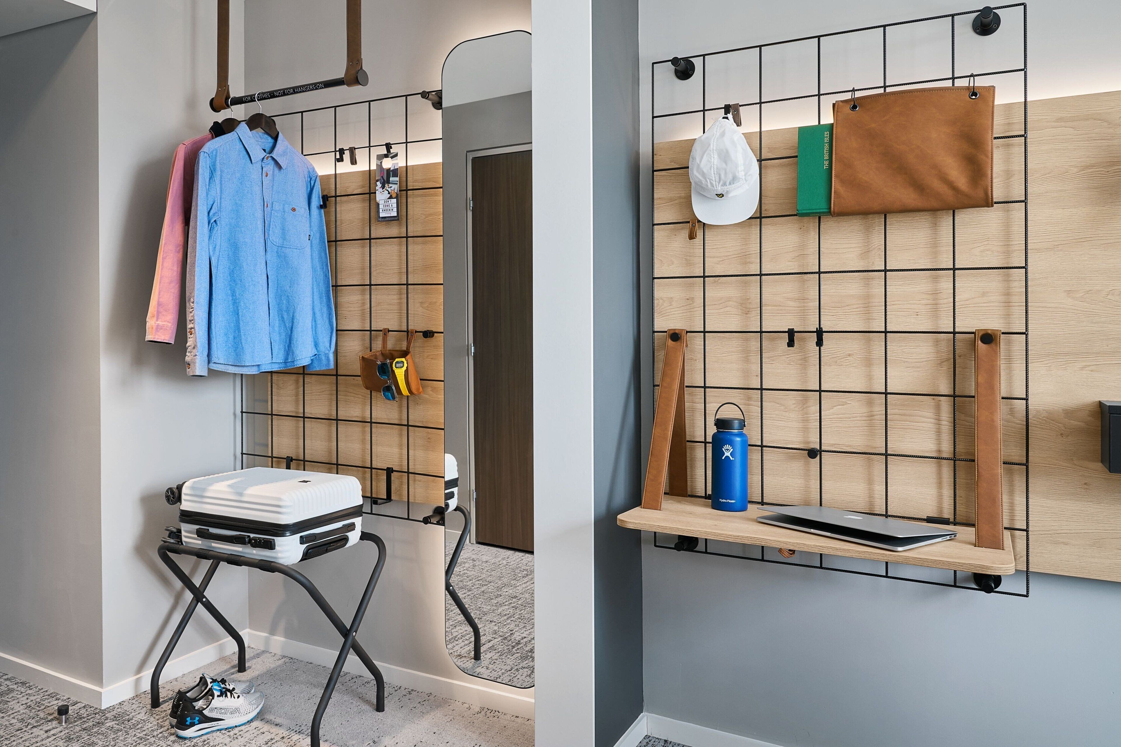 A room with a luggage rack, a small table with a laptop, and a wall-mounted shelf with a water bottle and a cap.
