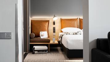 A compact hotel room with a bed, a small seating area with a pillow and a laptop, a bedside table with a lamp, and a power outlet.