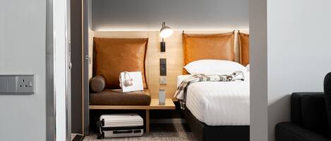 A compact hotel room with a bed, a small seating area with a pillow and a laptop, a bedside table with a lamp, and a power outlet.