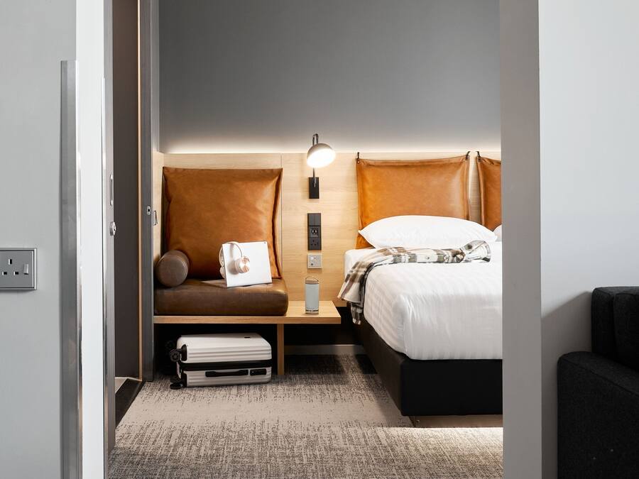 A compact hotel room with a bed, a small seating area with a pillow and a laptop, a bedside table with a lamp, and a power outlet.