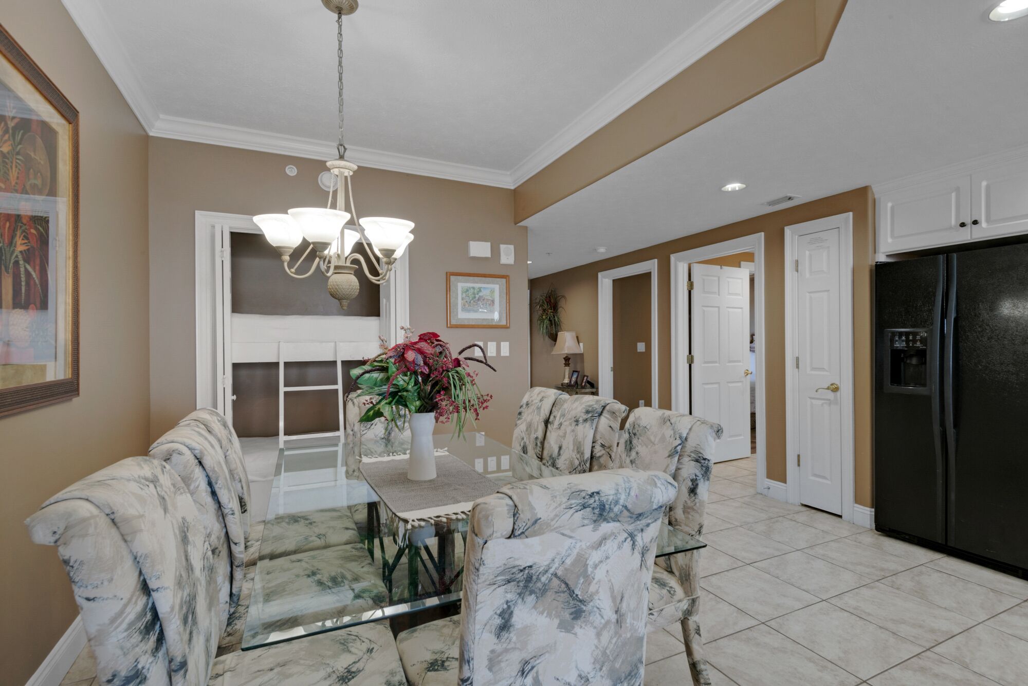 A dining area with a glass-top table, floral chairs, a chandelier, and a kitchen with a black refrigerator.