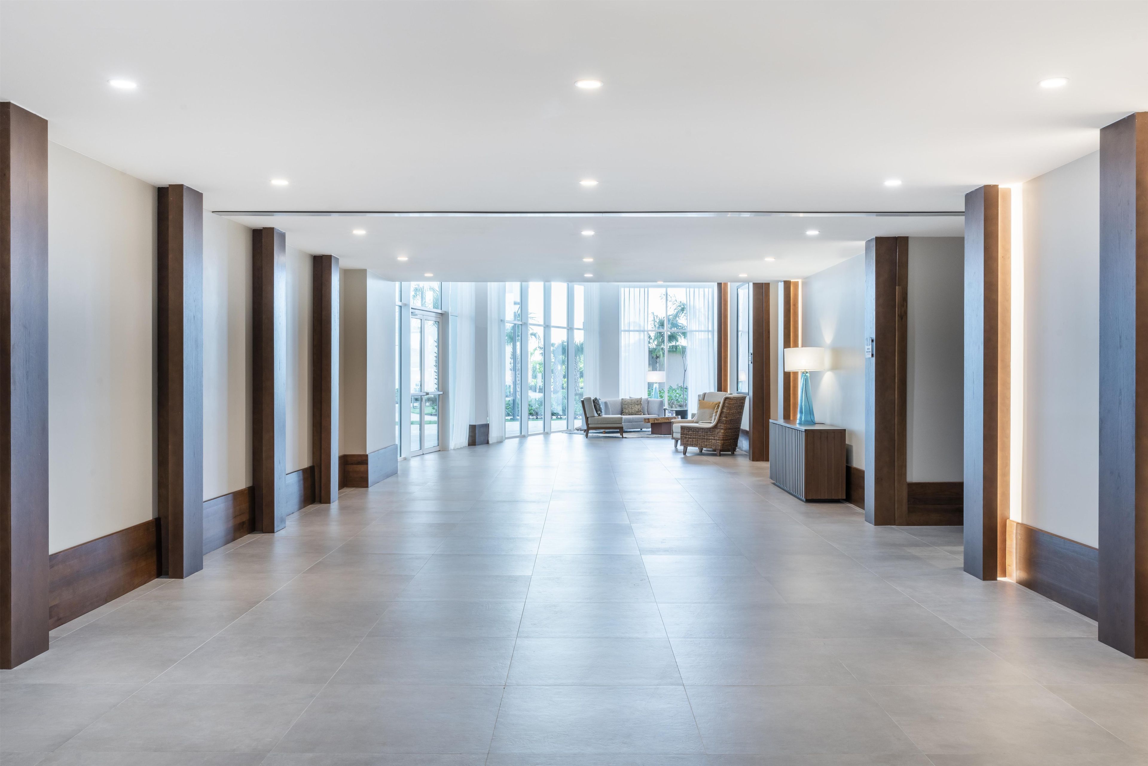 A spacious hotel lobby with a modern design, featuring a long corridor, seating area, and large windows.