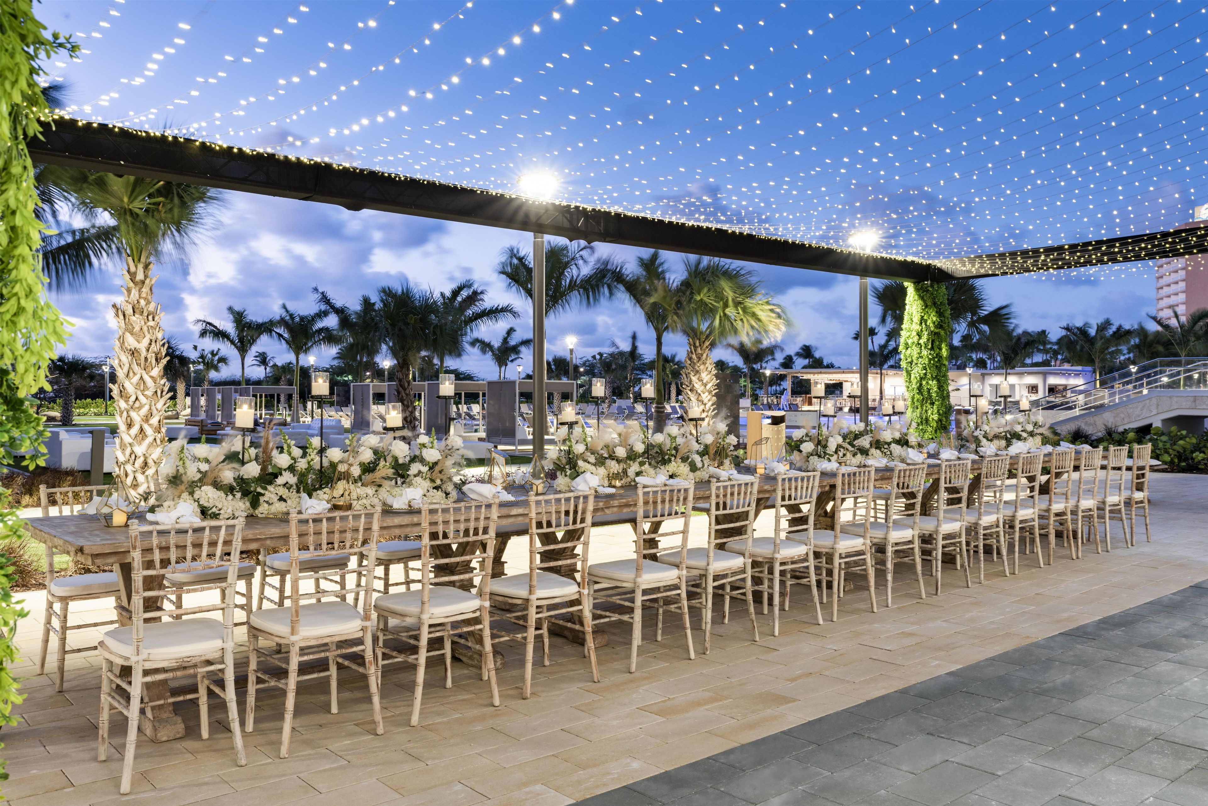 An outdoor dining area with multiple tables and chairs, palm trees, and string lights.
