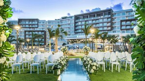 A hotel with a modern design, featuring a central pathway with white chairs and floral arrangements, surrounded by palm trees and a pool area.