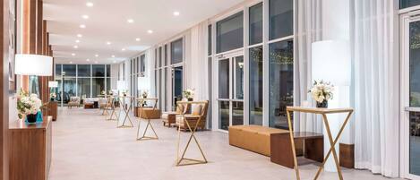 A spacious hotel lobby with a long reception desk, multiple seating areas, and large windows providing natural light.