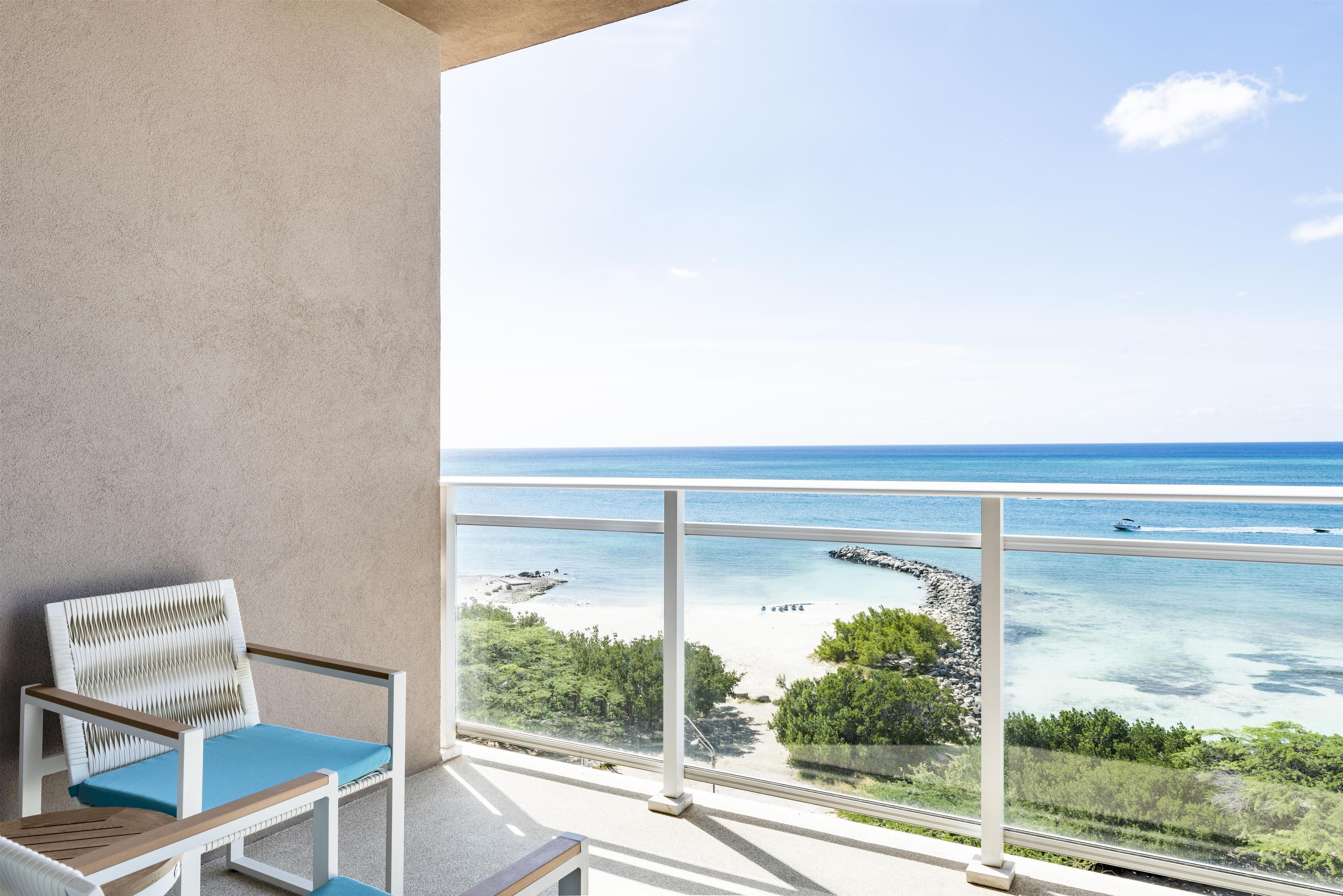 A balcony with a view of a beach, a chair, and a railing.