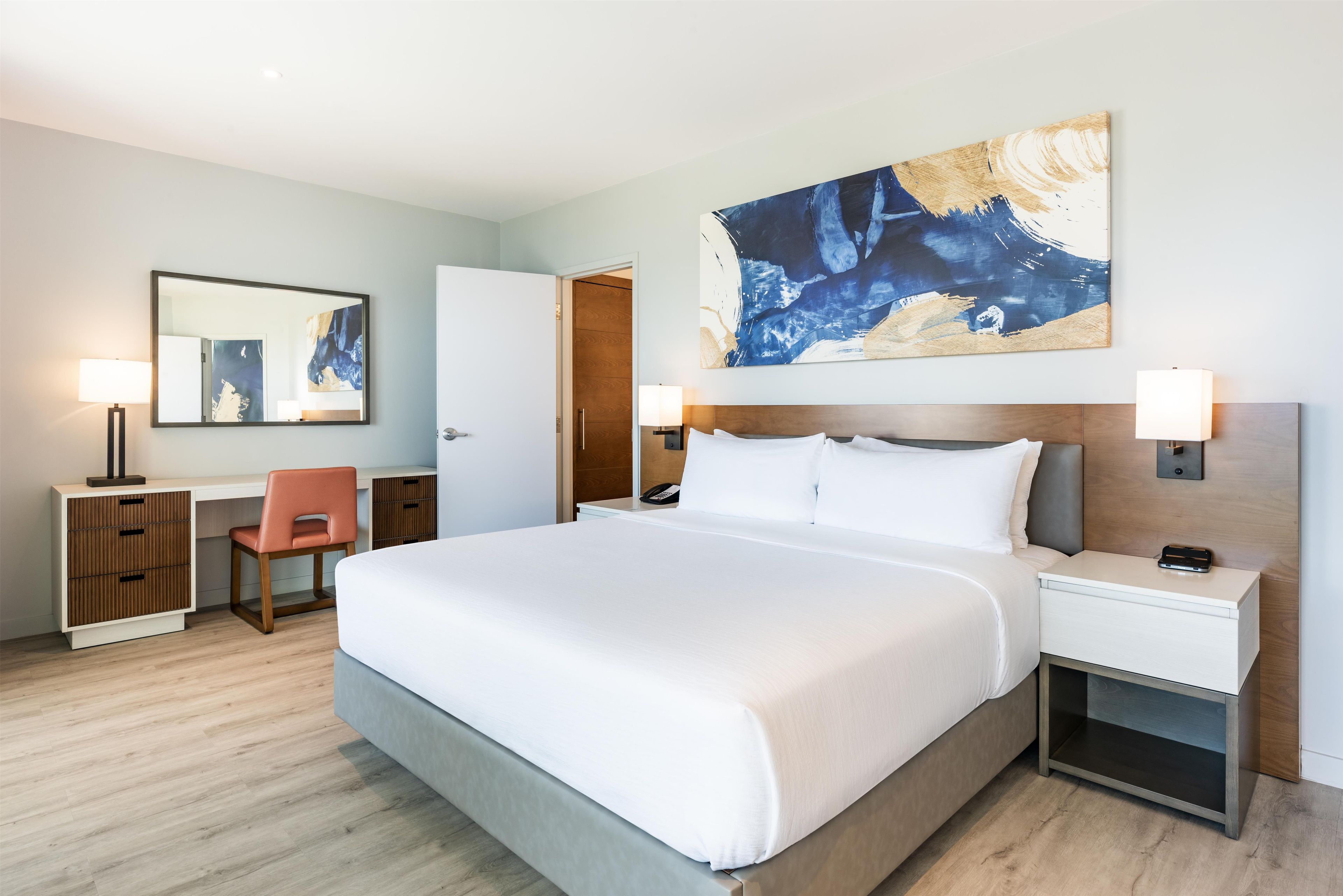 Une chambre d’hôtel avec un grand lit, deux tables de chevet, un bureau avec une chaise et un grand tableau abstrait accroché au mur.