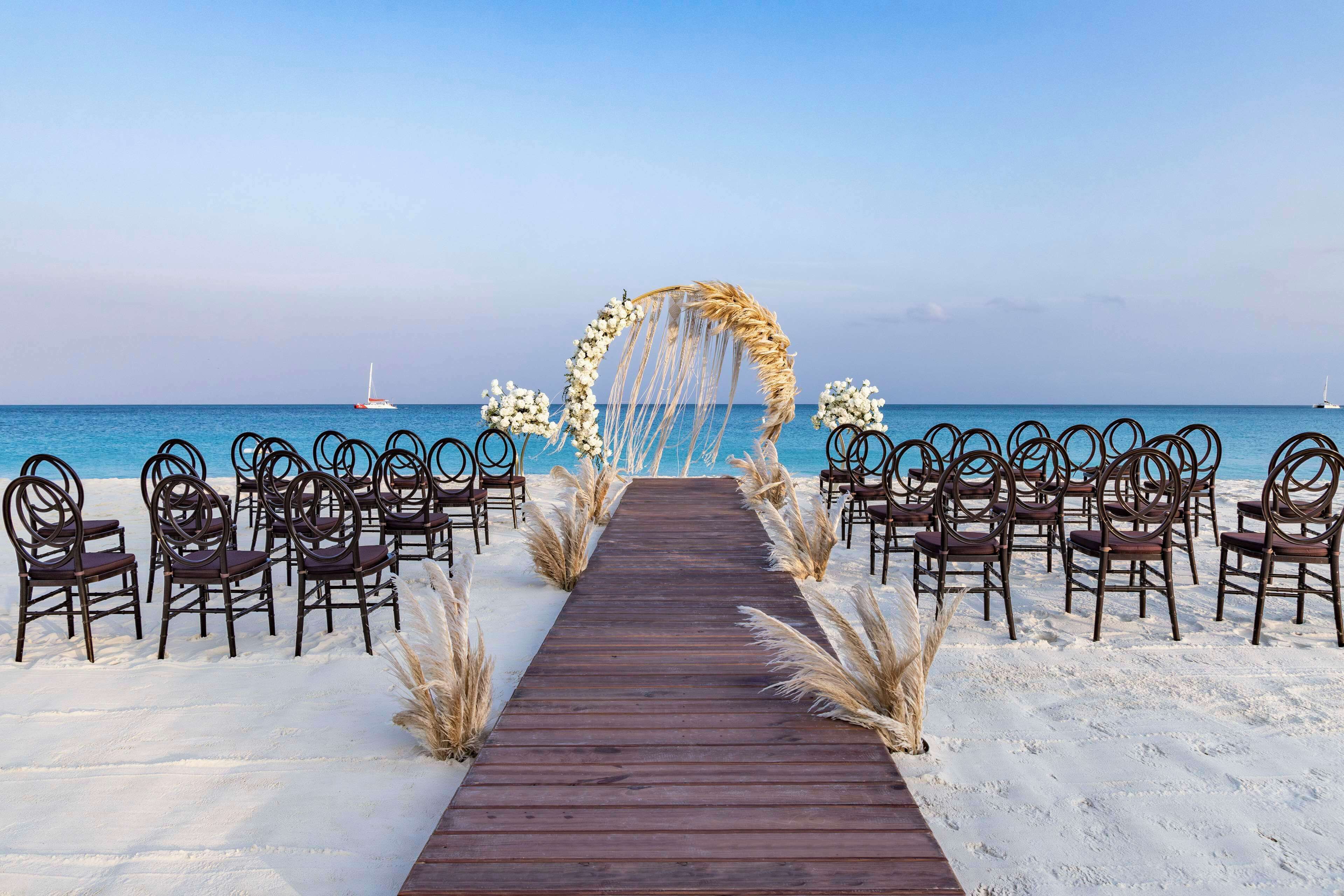 A wooden pathway leading to a beach wedding arch with chairs on either side.