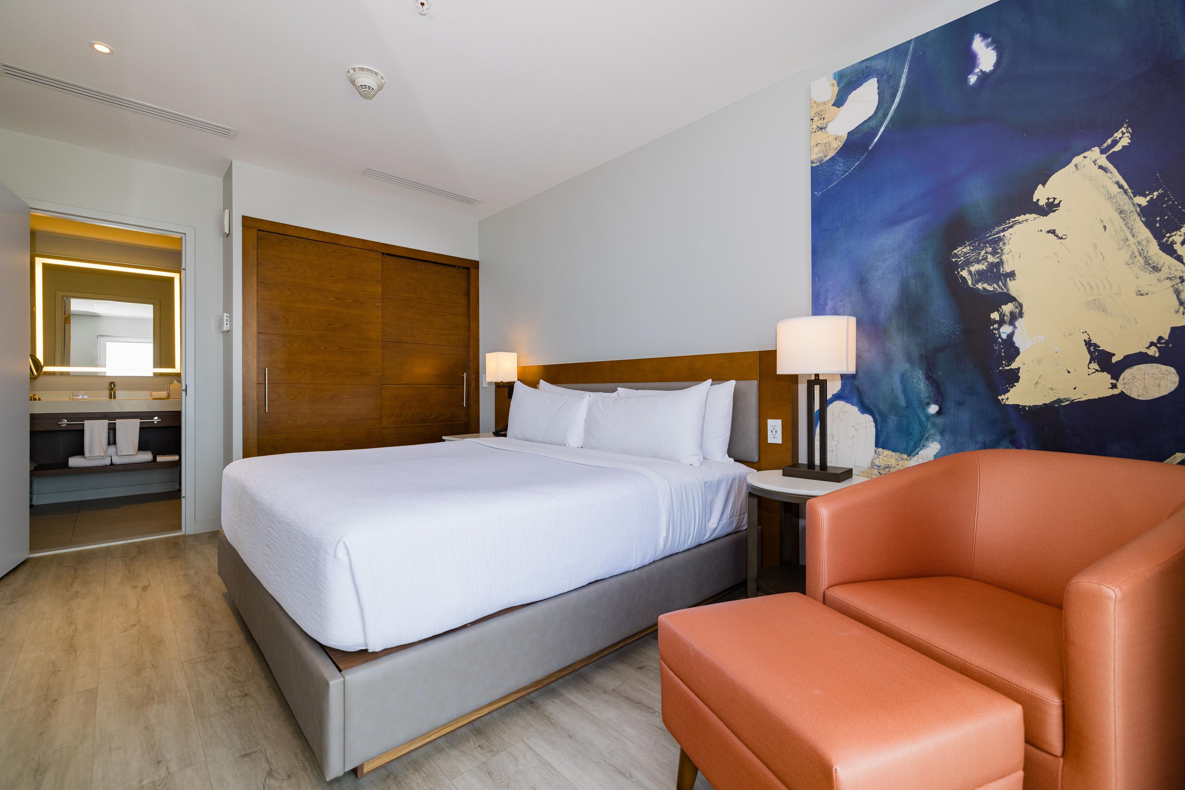 Habitación de hotel con cama, una silla, baño con espejo y un cuadro abstracto grande en la pared.
