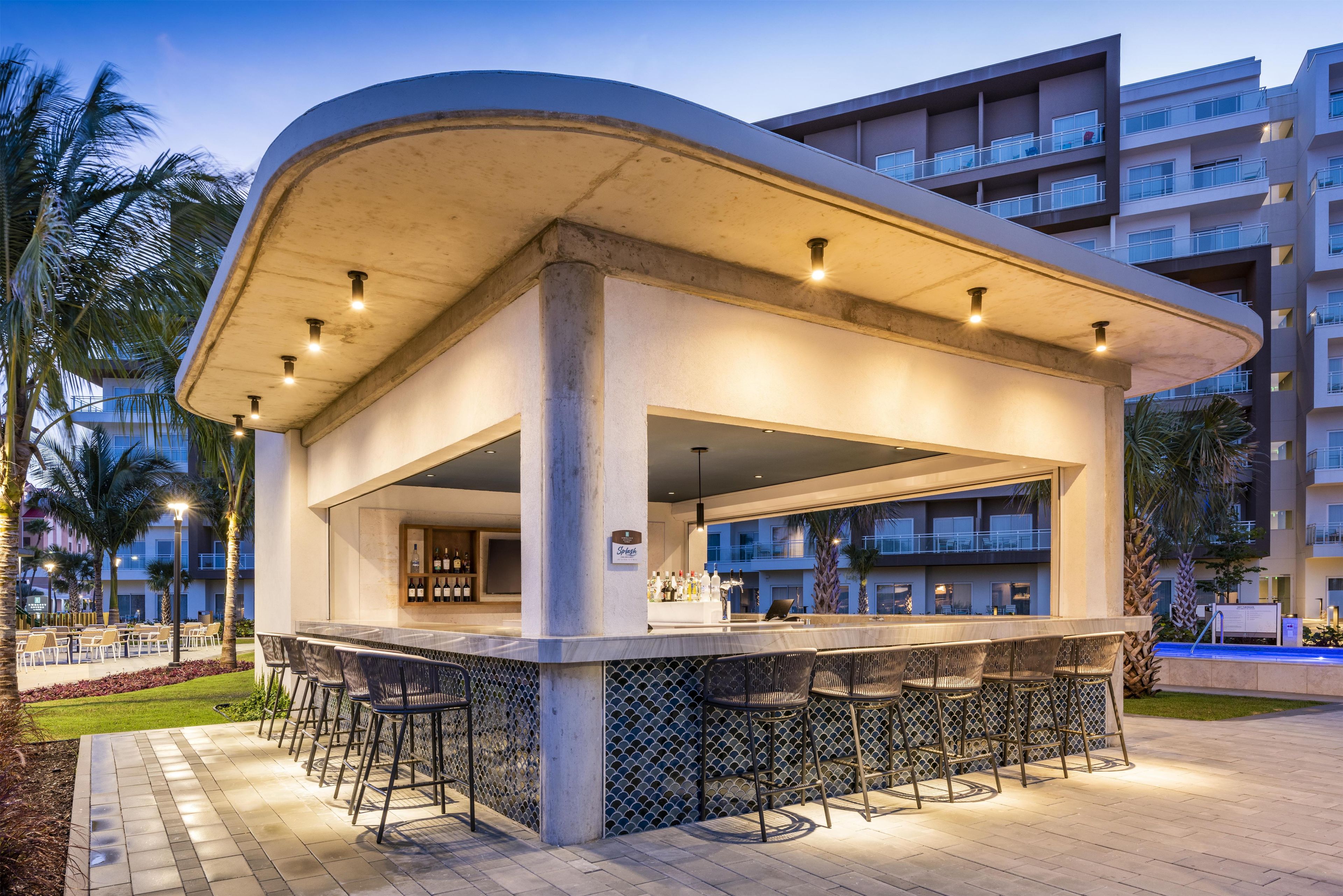 Un área de bar al aire libre con techo curvo, diversas opciones de asientos y vista a un edificio moderno al fondo.