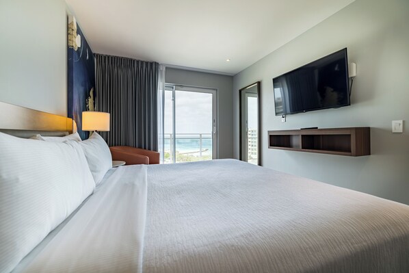 Una habitación de hotel con una cama grande, un televisor montado en la pared, una ventana con vistas al mar y una puerta corrediza que da a un balcón.