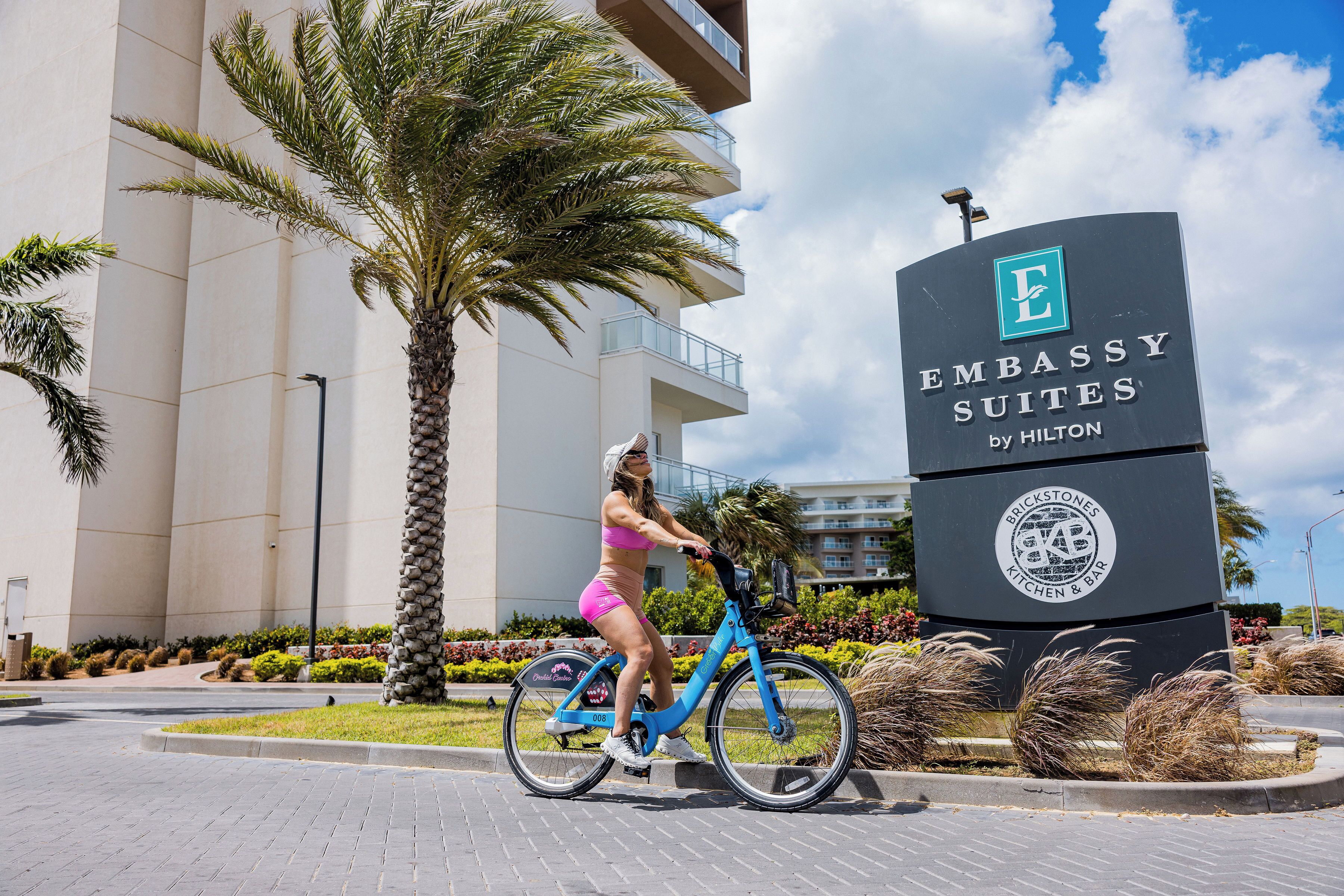 Una mujer pedalea una bicicleta eléctrica azul frente a un cartel de Embassy Suites by Hilton.